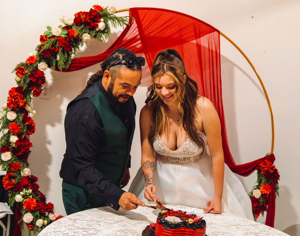 Couple cutting wedding cake under red floral arch with roses and red draping at elegant wedding ceremony.