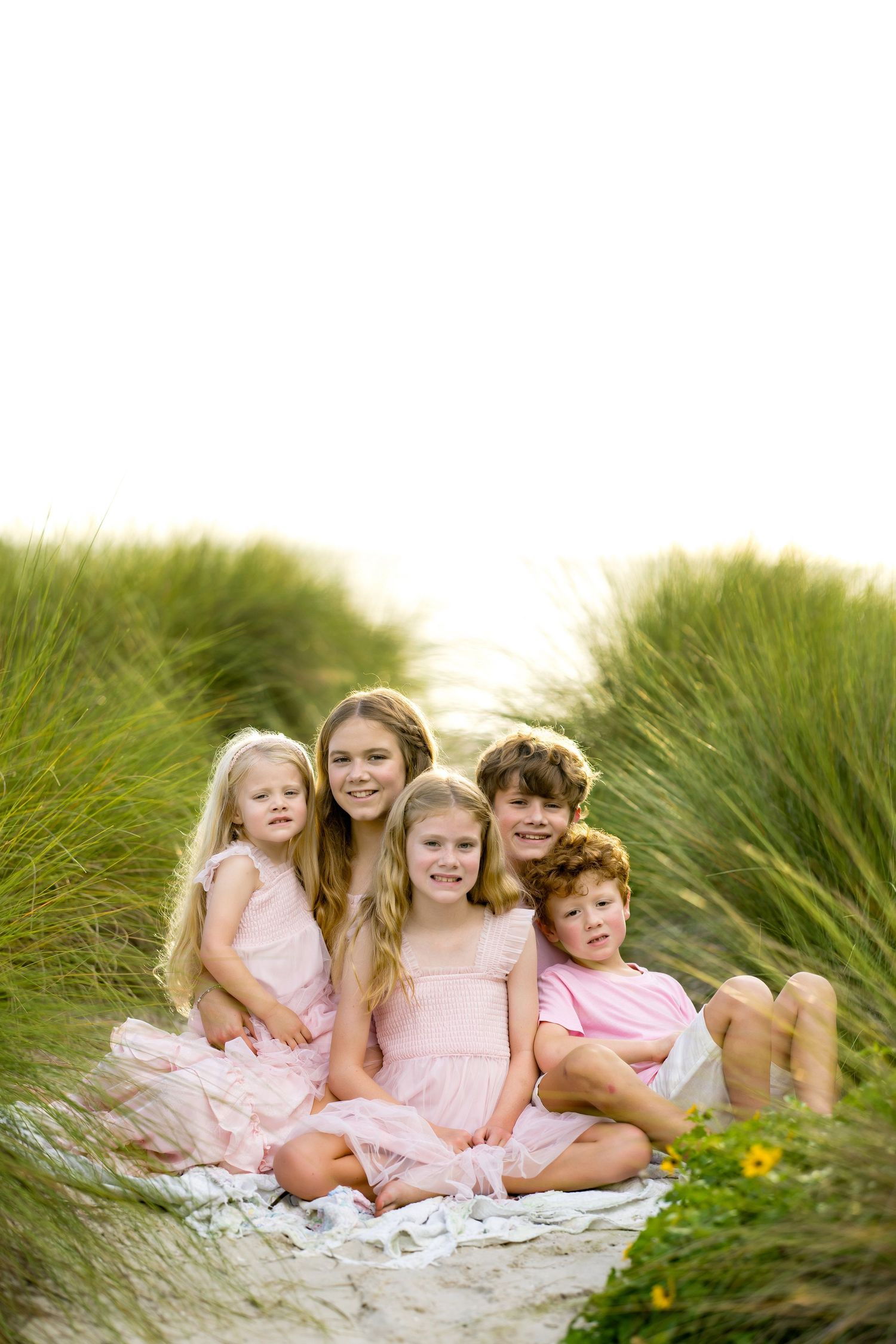 Five children in pink outfits sitting together on a sandy path surrounded by tall green grass.