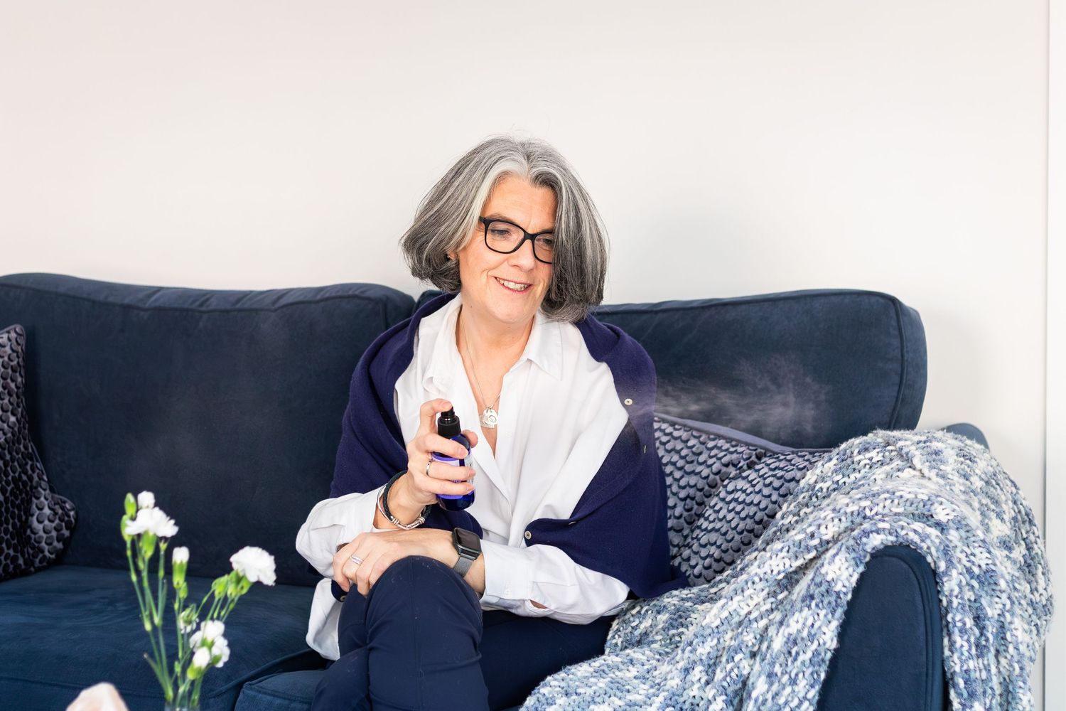 Smiling middle-aged woman with gray hair and glasses relaxing on a navy sofa holding a TV remote.