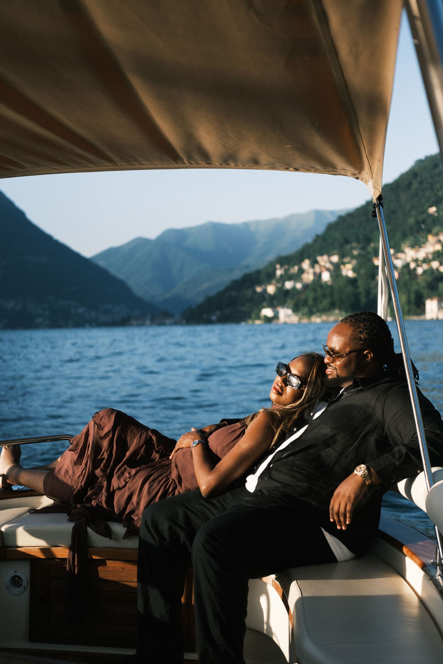 Elegant couple relaxing on a luxury yacht with stunning mountain lake views, woman in brown dress, man in black suit.