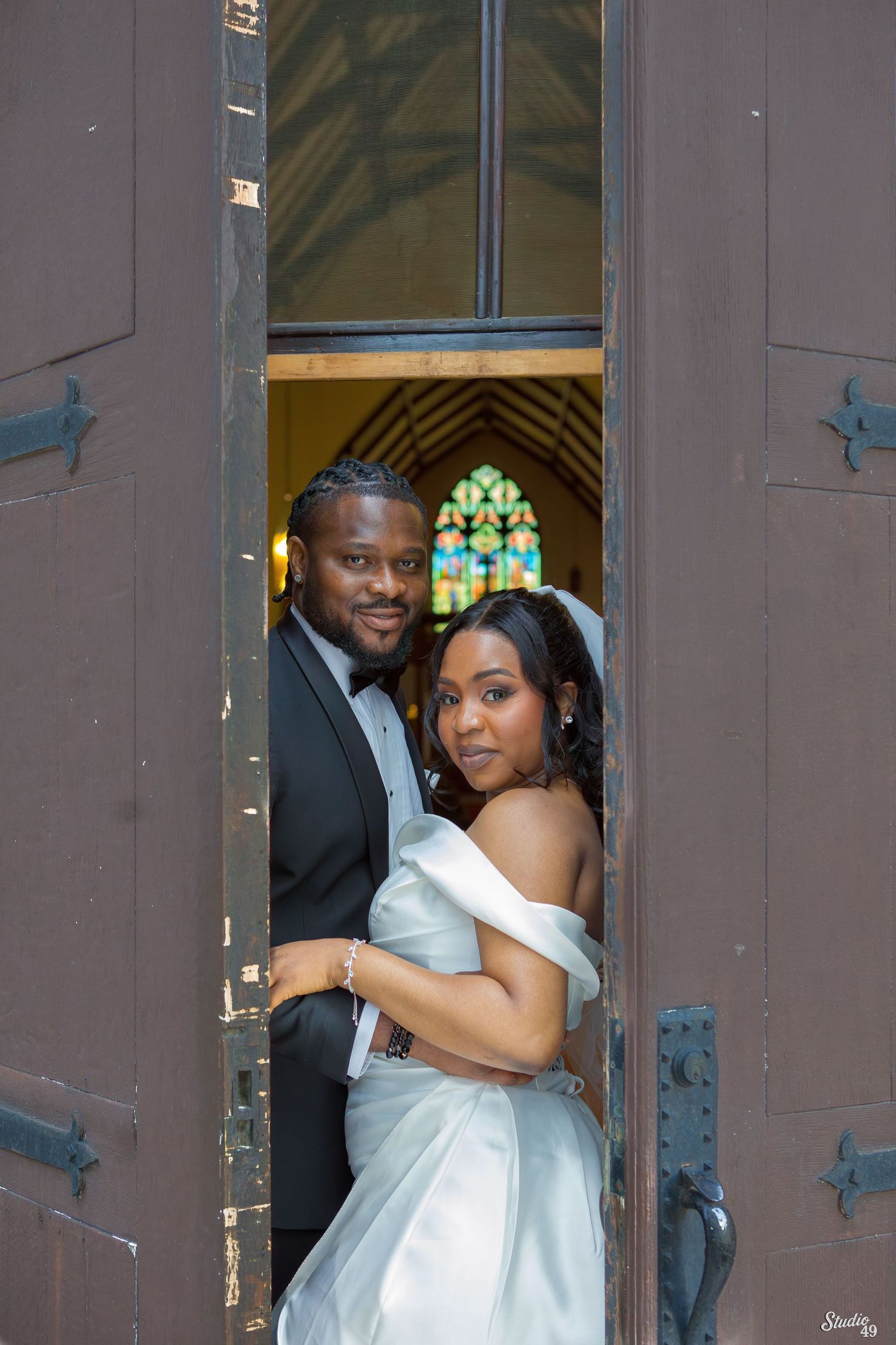 Bride and groom posing in church doorway with stained glass window behind them.
