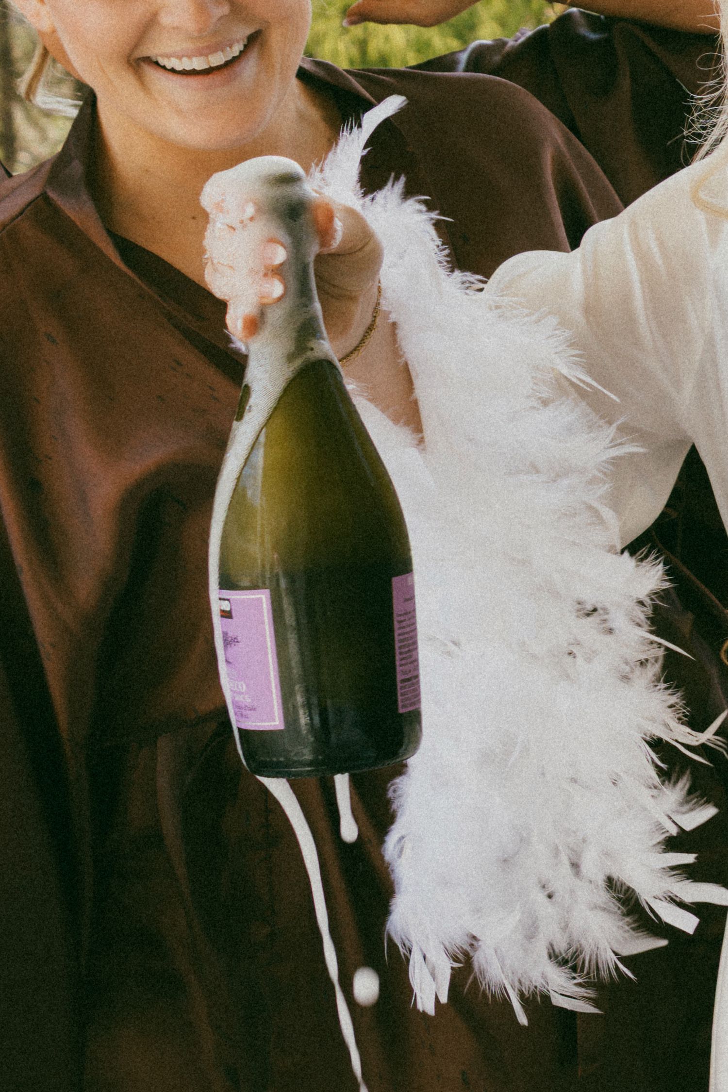 Champagne bottle overflowing with foam held by two people, one wearing a white feather boa, celebrating outdoors.