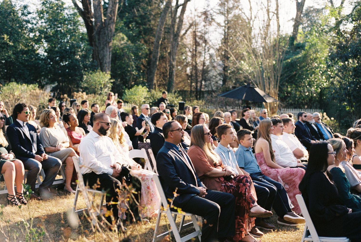 Guests seated outdoors at a garden wedding ceremony on white chairs, surrounded by lush trees on a sunny day.