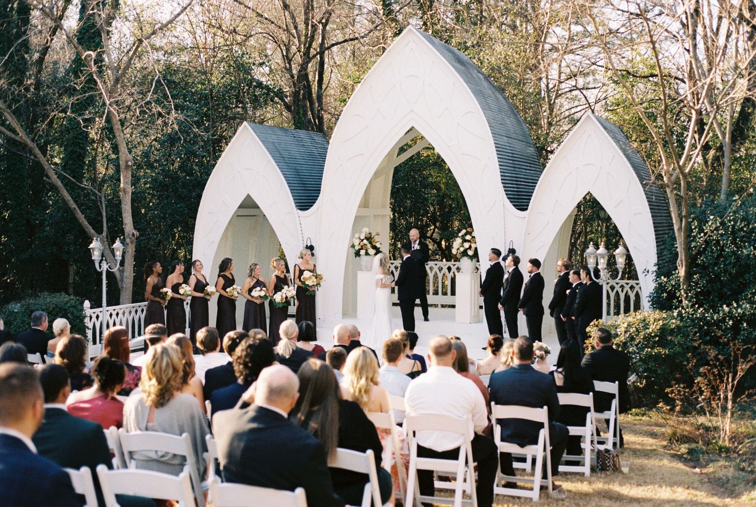Outdoor wedding ceremony in Febuary with guests seated before a white arch backdrop surrounded by trees.