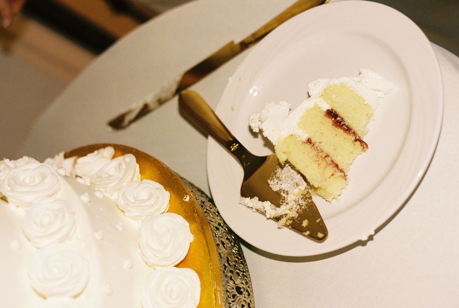 A slice of white wedding cake with strawberry filling on a plate next to the decorated layered cake with rosette frosting.
