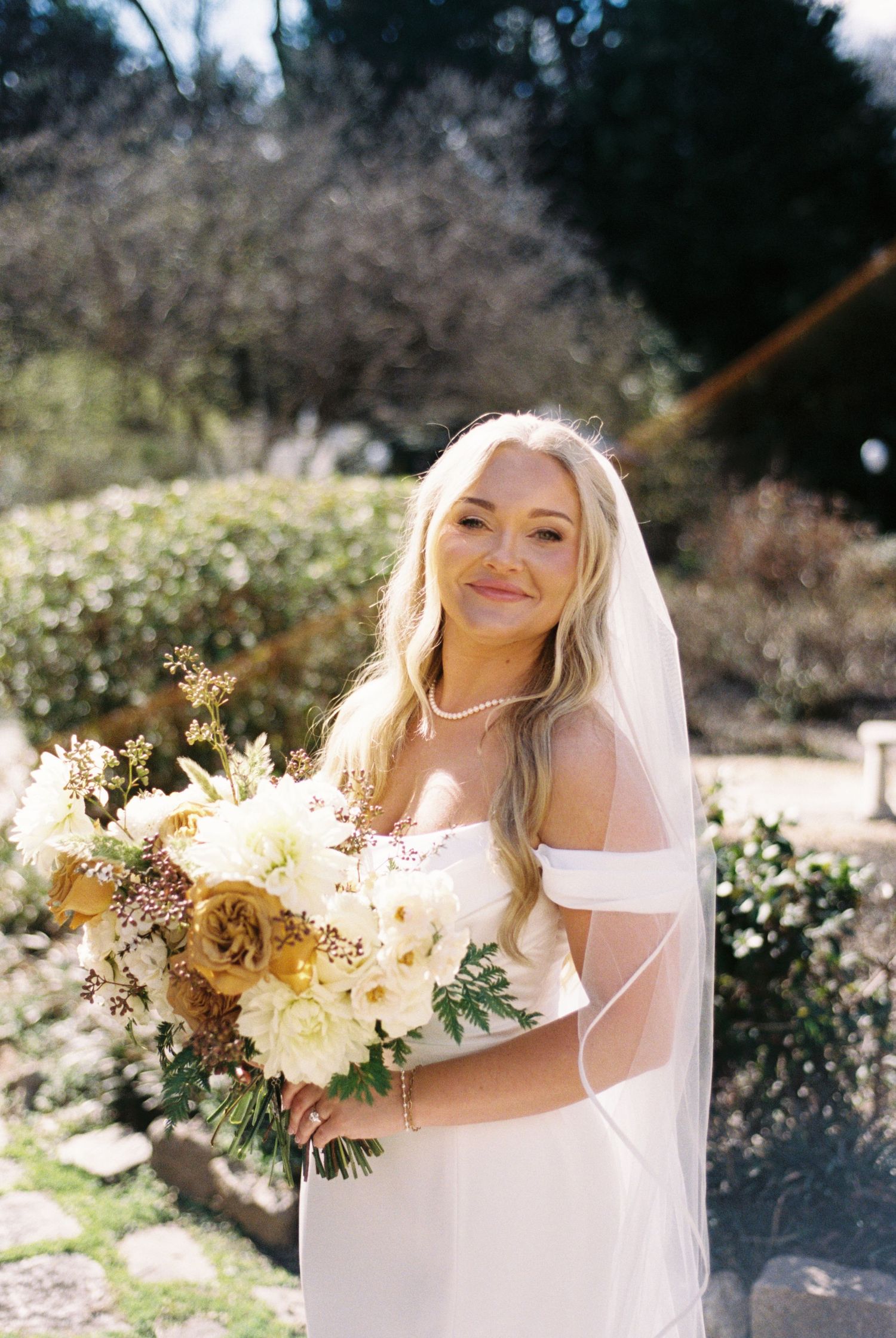 Smiling bride in off-shoulder white gown and veil holds yellow floral bouquet outdoors on sunny day.