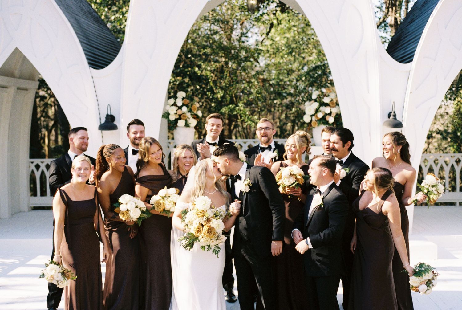 Wedding party in black and brown attire poses under white arch with bride and groom kissing at outdoor ceremony, shot with 35mm film.