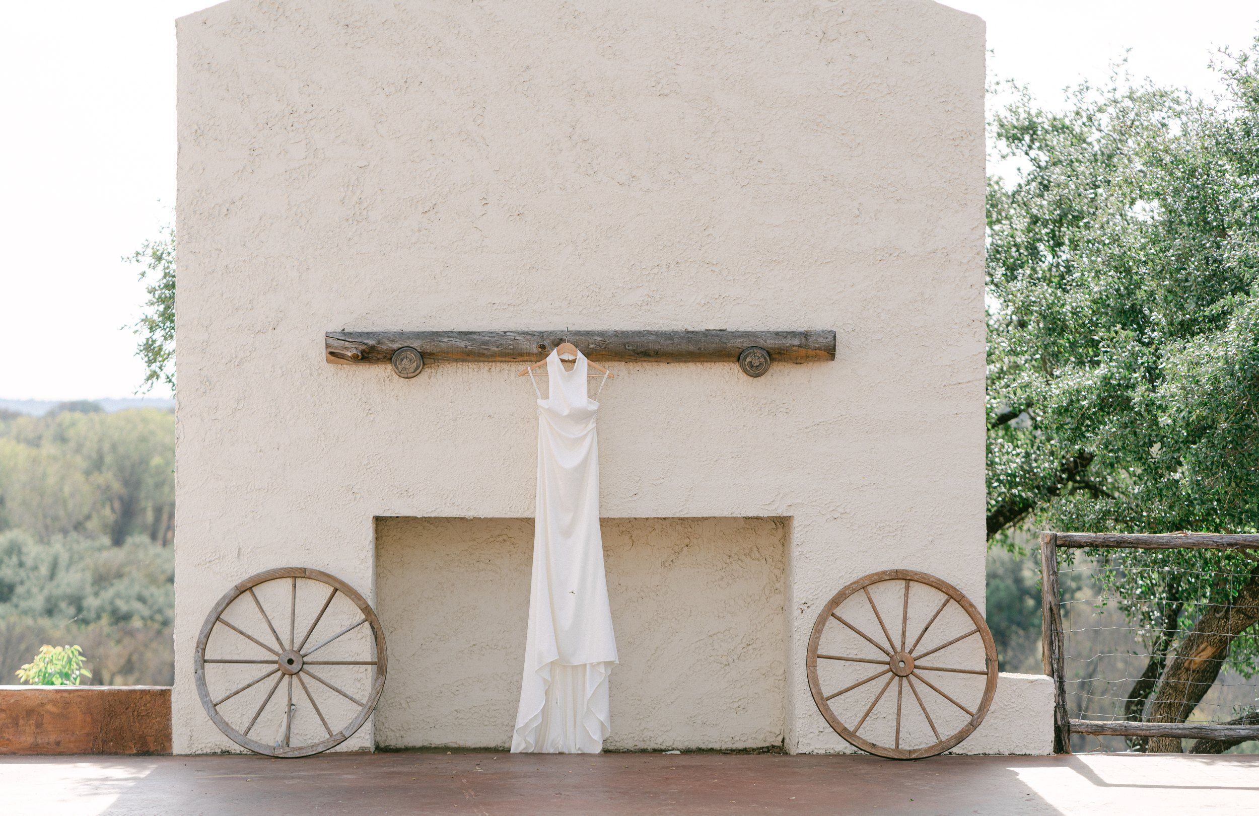 A white halter wedding gown hanging from a rustic beam on a plaster wall between two wagon wheels, Texas Hill Country