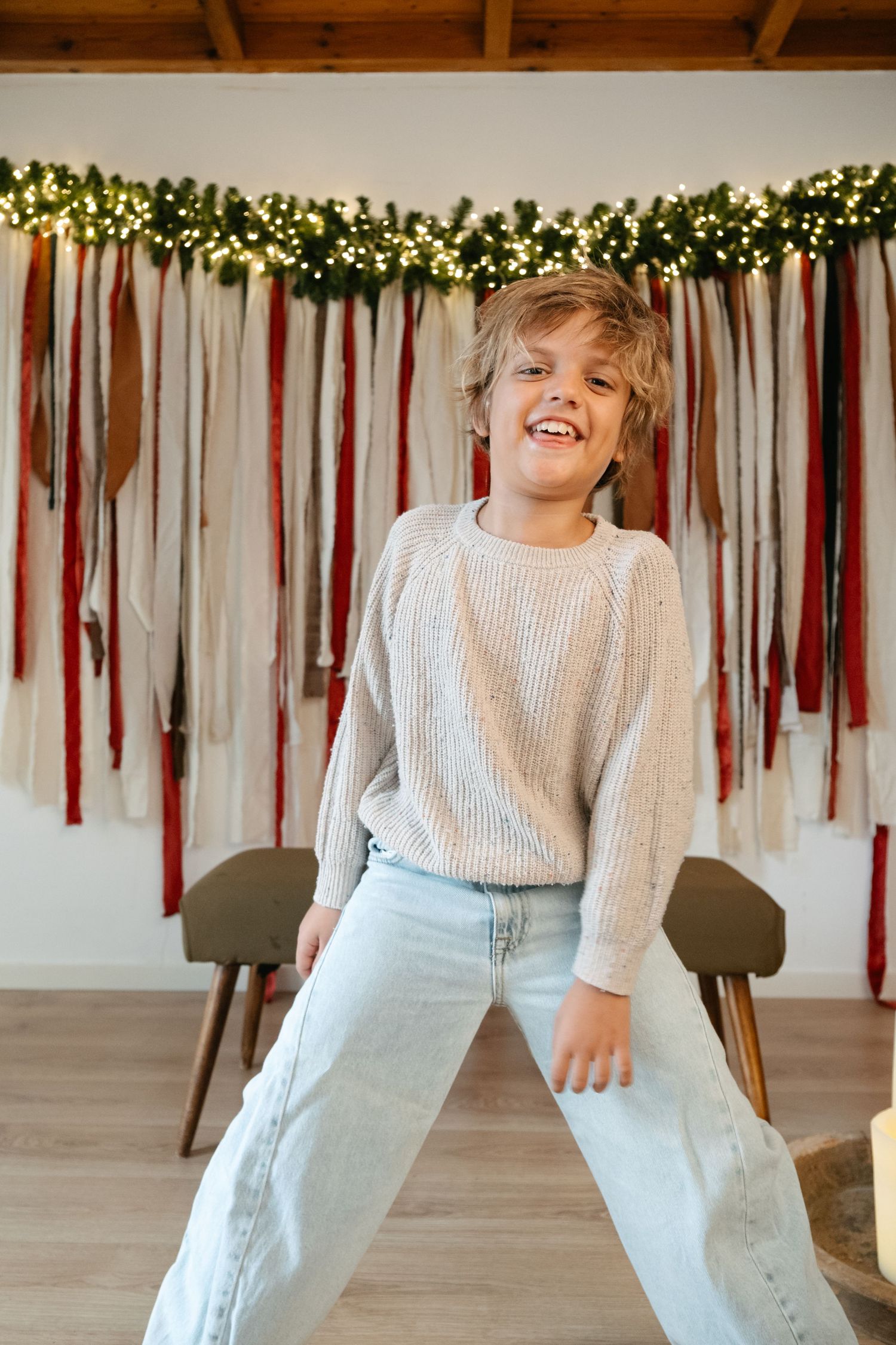 Smiling boy in a cream knit sweater and light jeans stands with legs apart in front of a festive ribbon and string light garland backdrop.