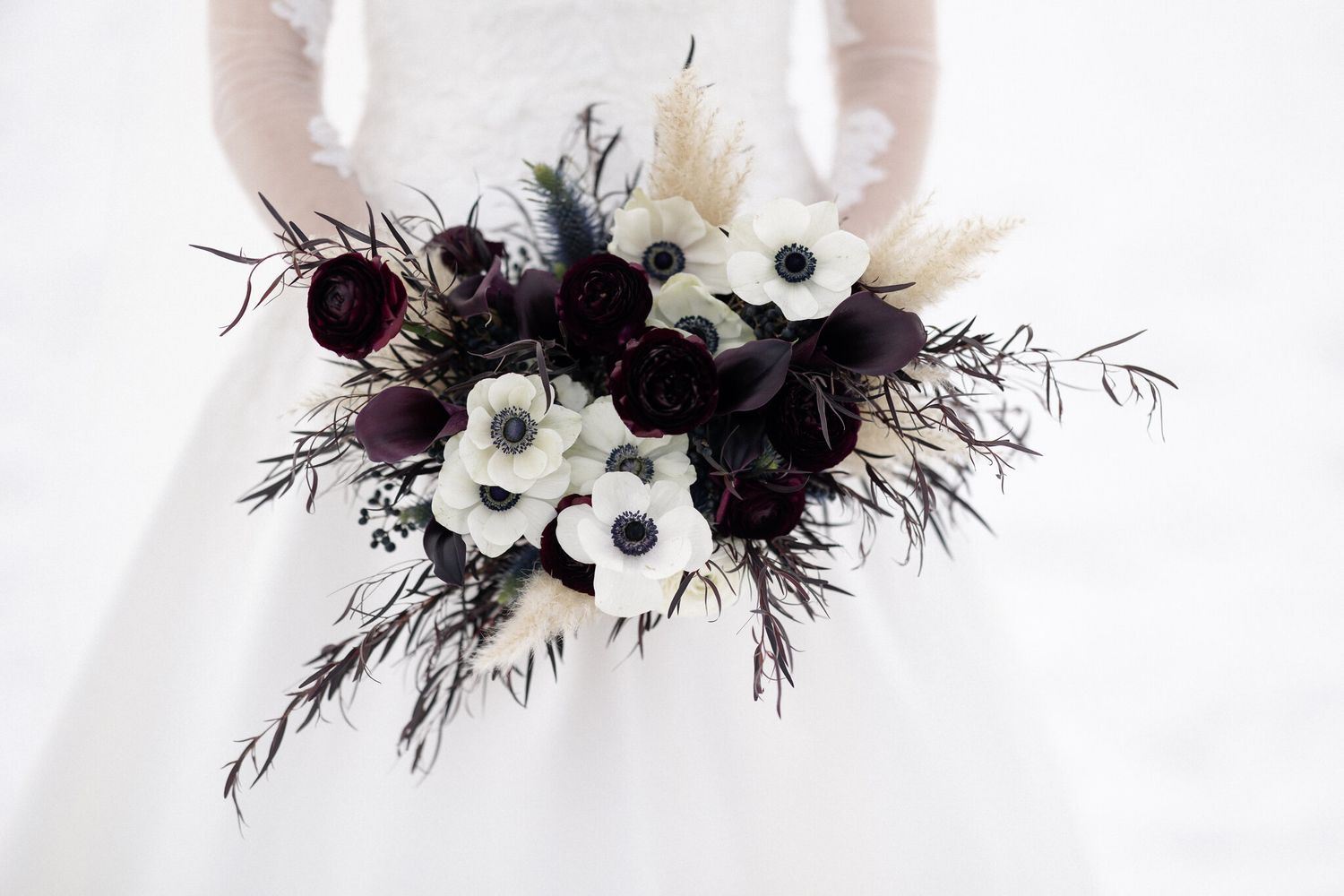 Winter wedding bouquet of white anemones, burgundy blooms and pampas against a lace gown in Banff