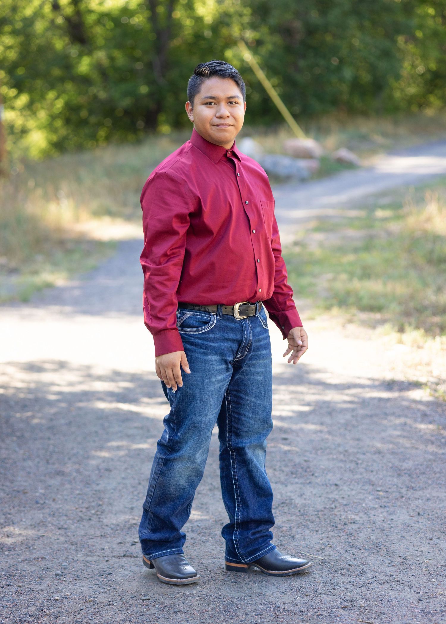 Young man in red shirt, jeans, and cowboy boots standing on a dirt path surrounded by trees.