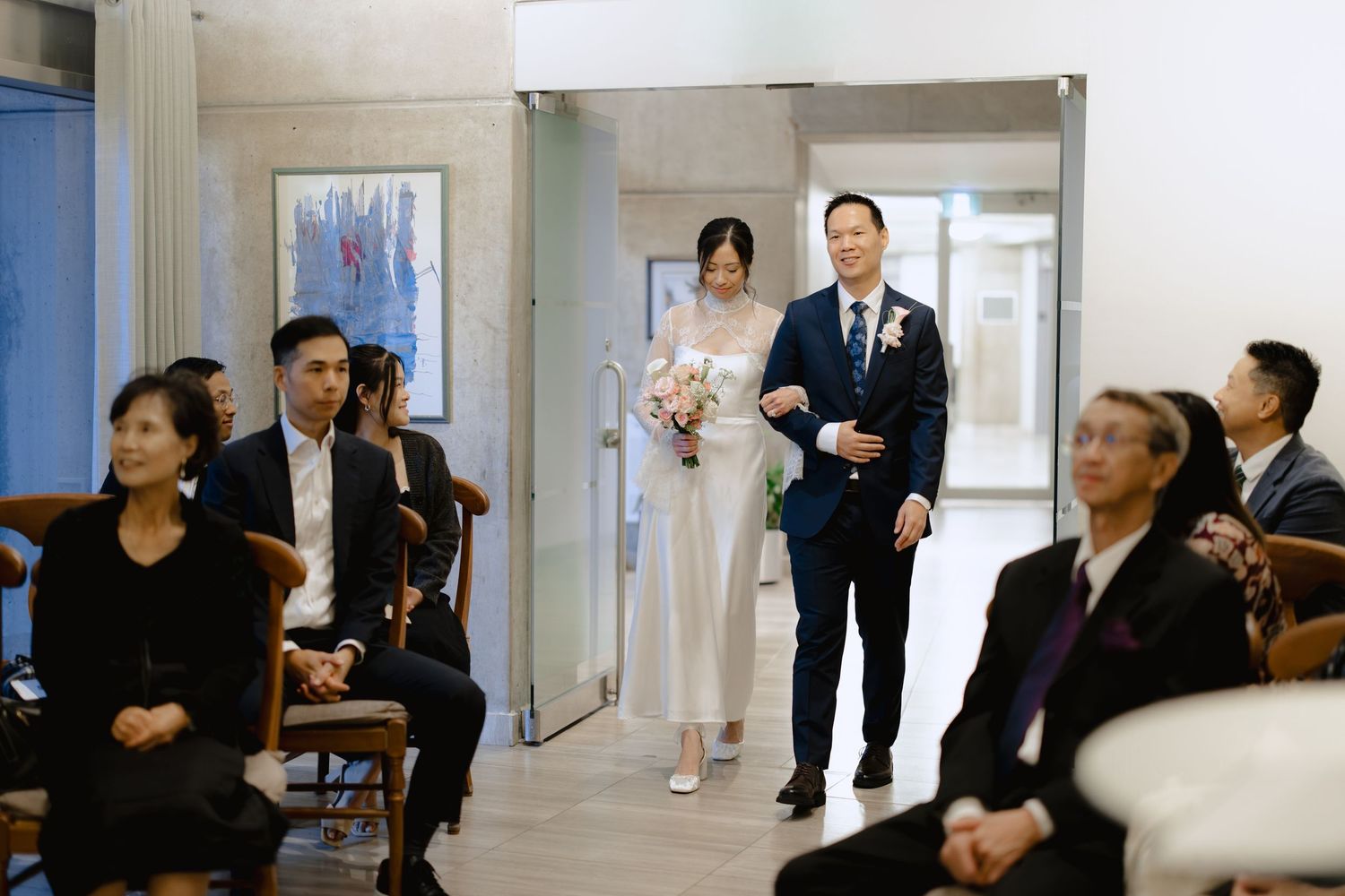 A bride in a white gown holding a bouquet walks down the aisle beside a man in a navy suit as seated wedding guests look on.