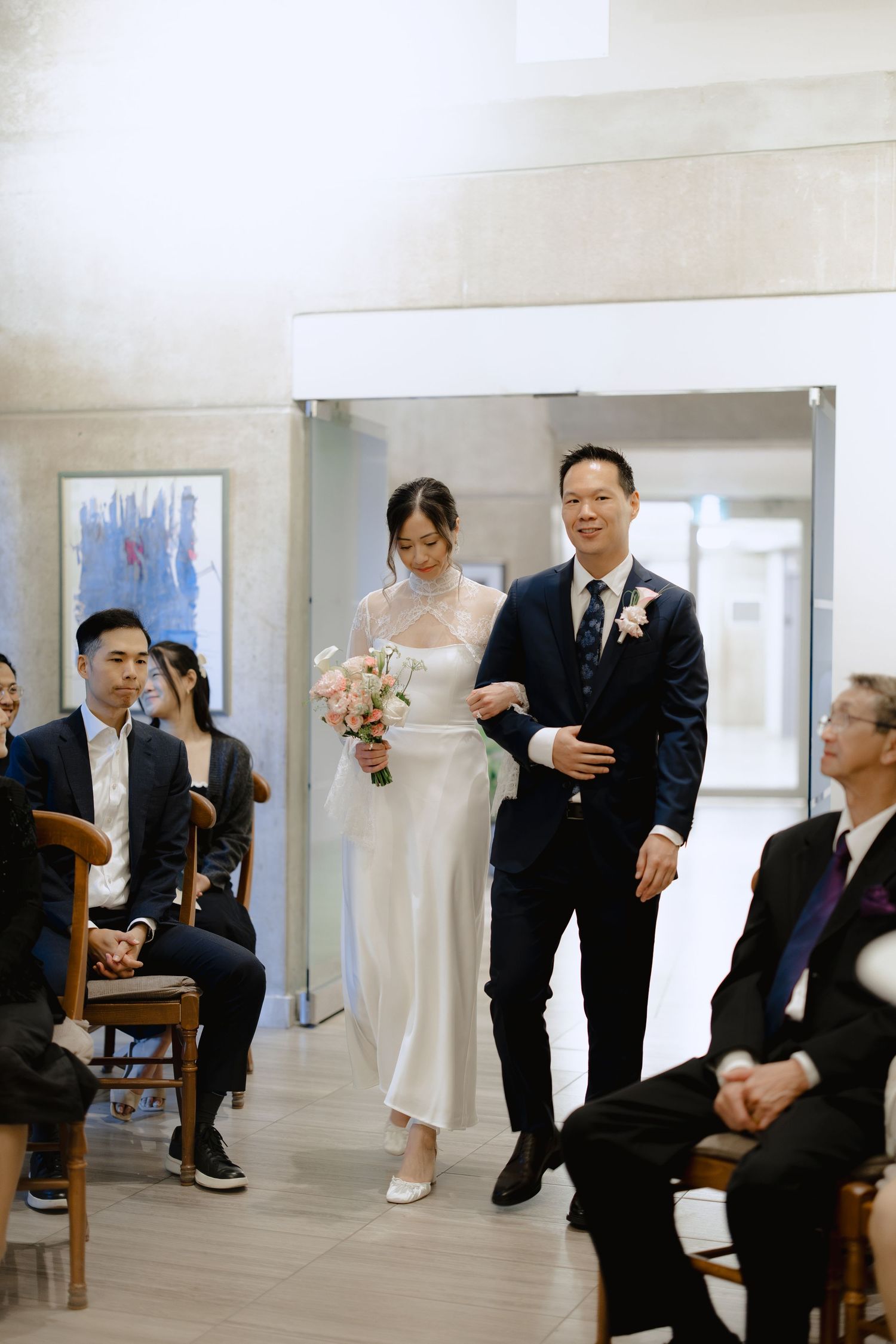 A bride in a white gown holding a pink bouquet walks down the aisle on the arm of a man in a navy suit as seated wedding guests look on.