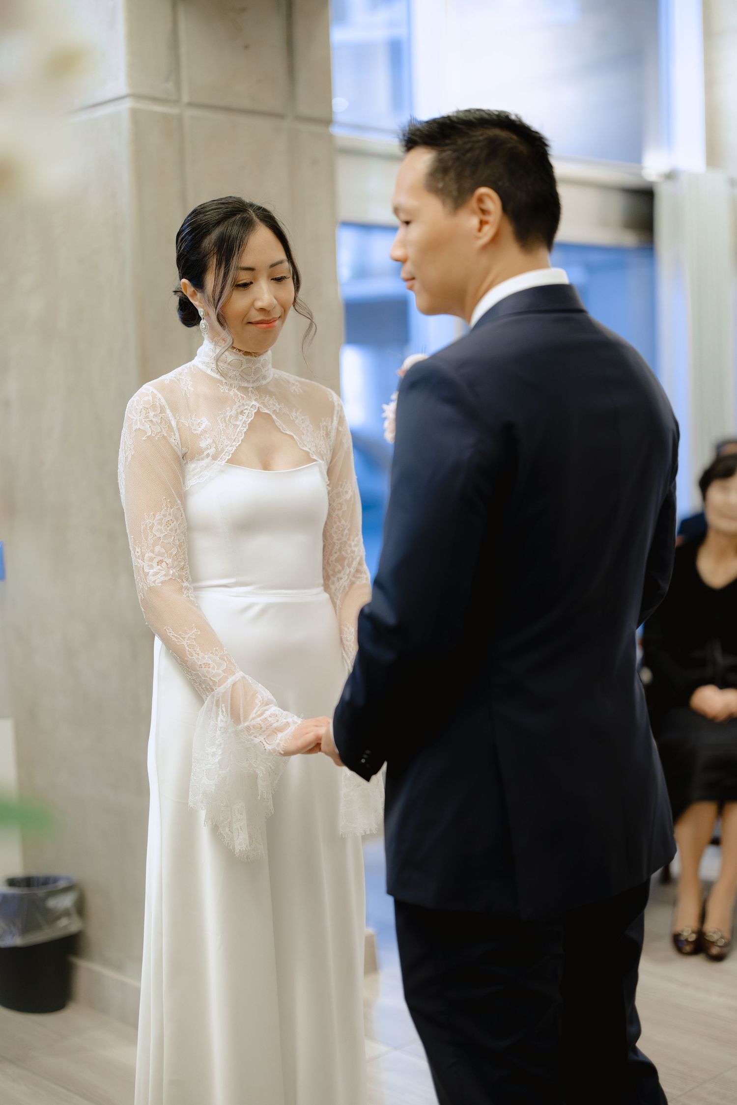 Bride in a white gown with sheer lace sleeves holds hands with the groom in a navy suit during their intimate wedding ceremony.