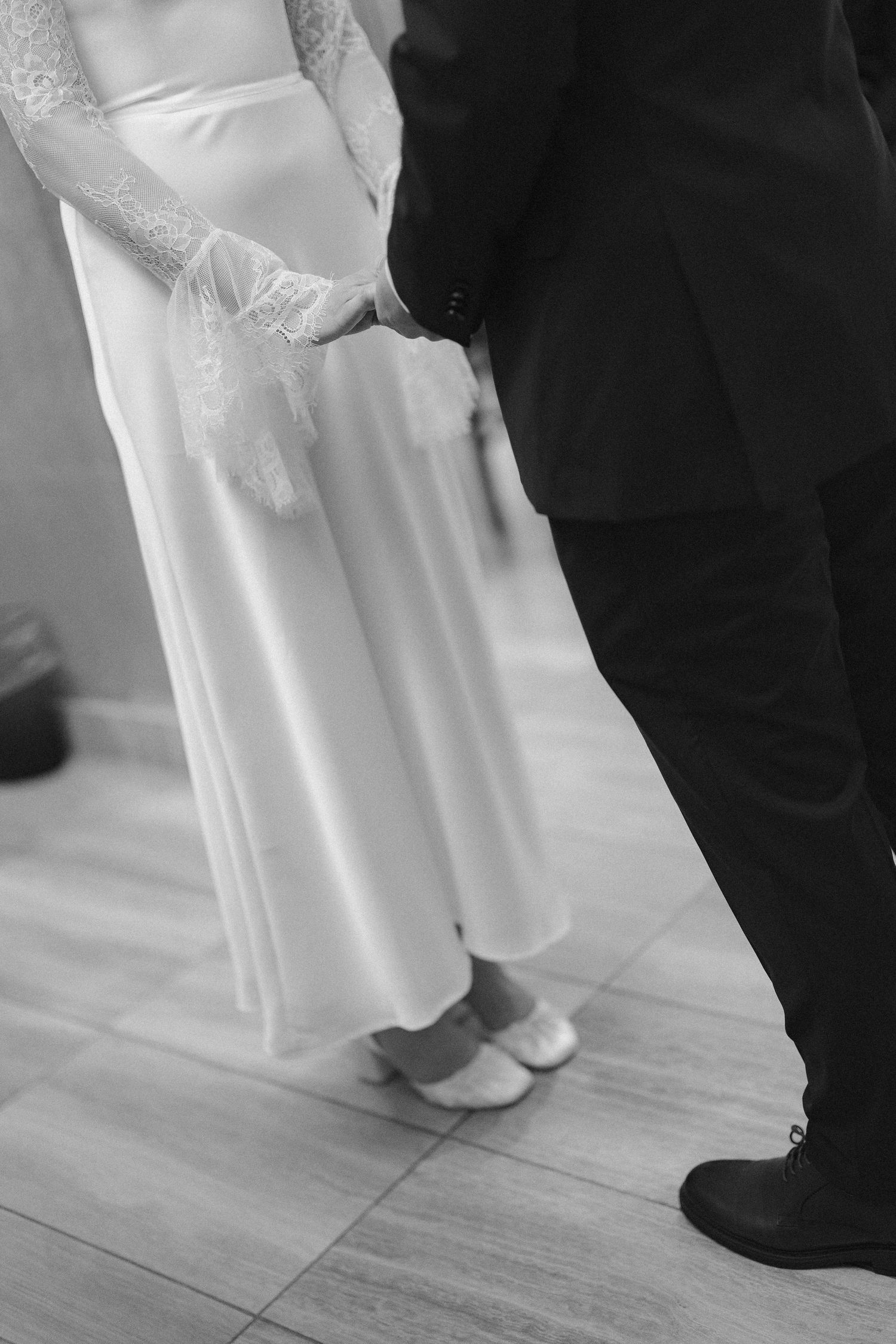 Black and white close-up of a bride in a lace-sleeved satin dress holding hands with a groom in a dark suit.