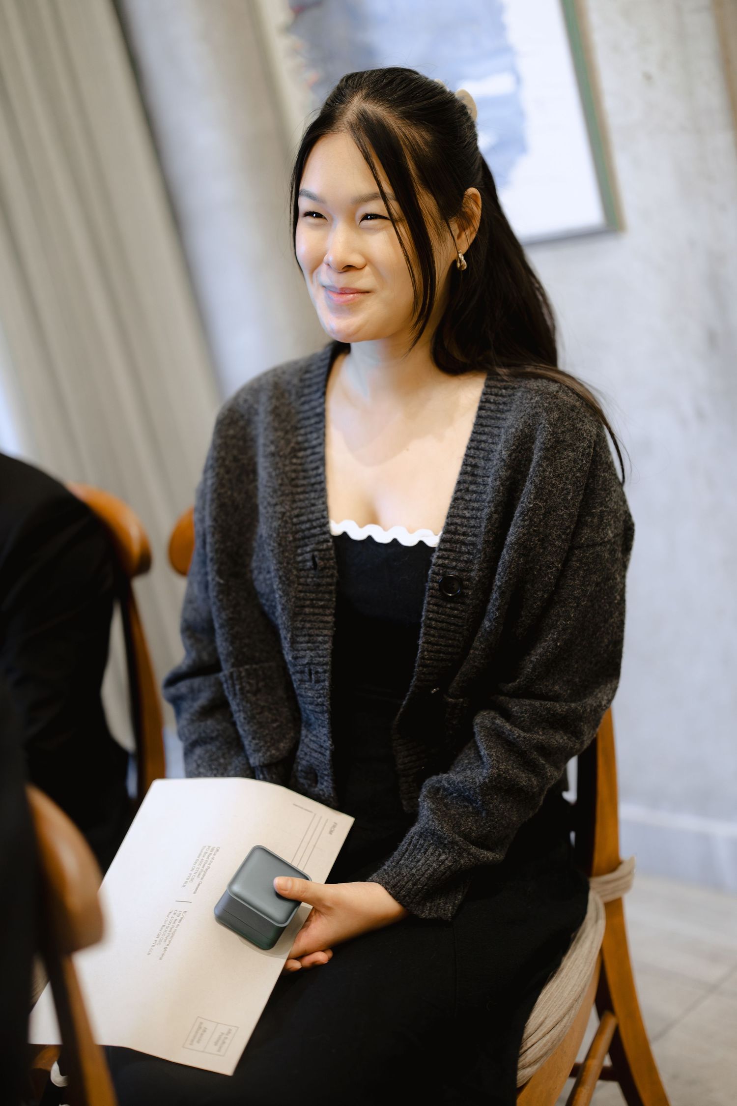 Smiling woman in a gray knit cardigan sits on a wooden chair holding a phone and envelope at an intimate wedding ceremony.