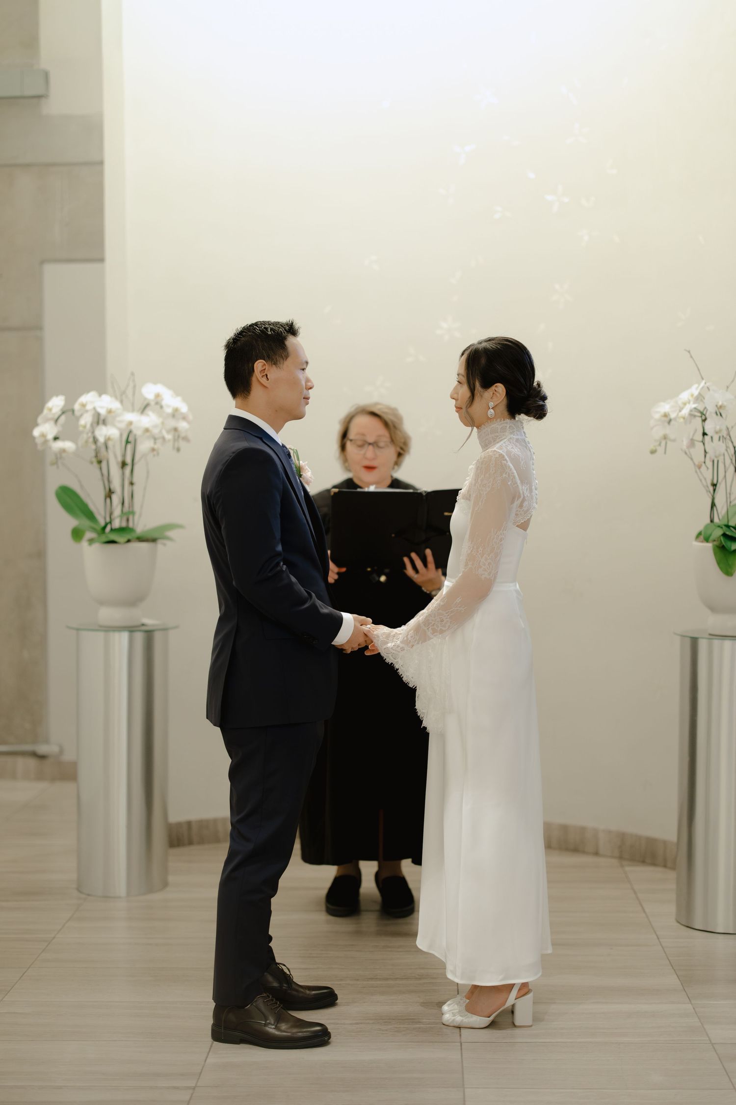 Bride and groom hold hands facing each other as an officiant reads from a black folder during their indoor ceremony.