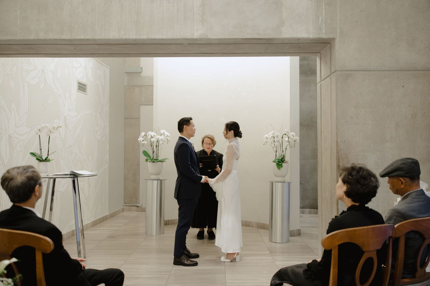 Bride in a white gown and groom in a dark suit hold hands before an officiant at a minimalist wedding ceremony with white orchids.