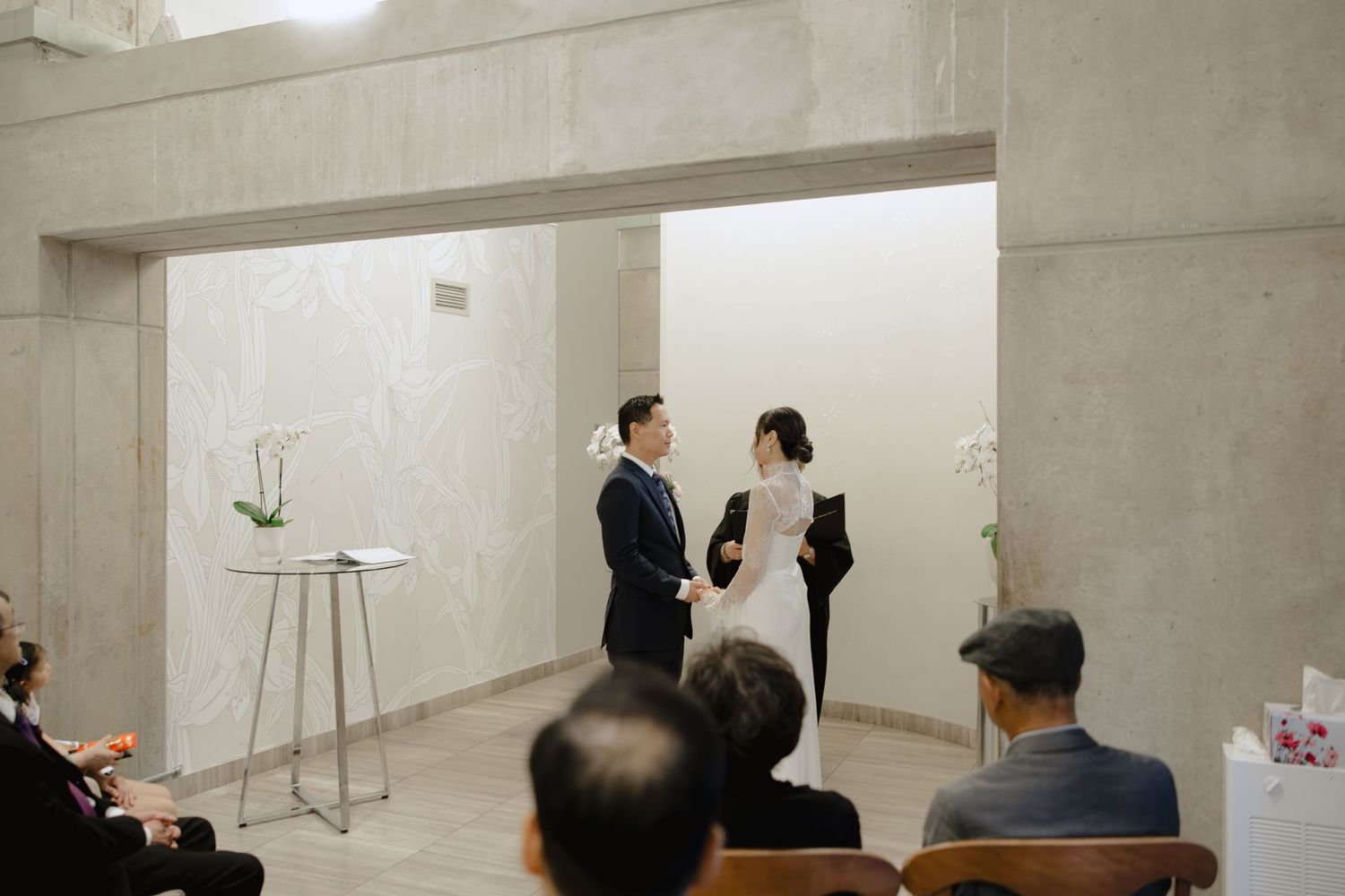 A couple stands facing each other during an intimate indoor wedding ceremony as seated guests watch in a modern, minimalist room.