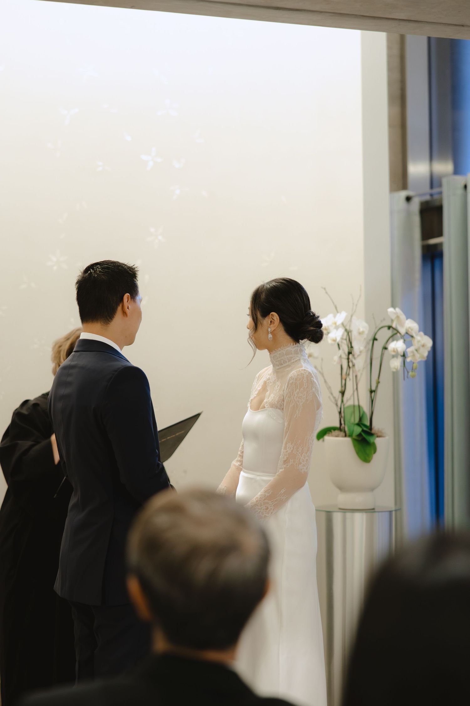 Bride in a white lace-sleeved gown and groom in a navy suit exchange vows at an intimate wedding ceremony as guests look on.
