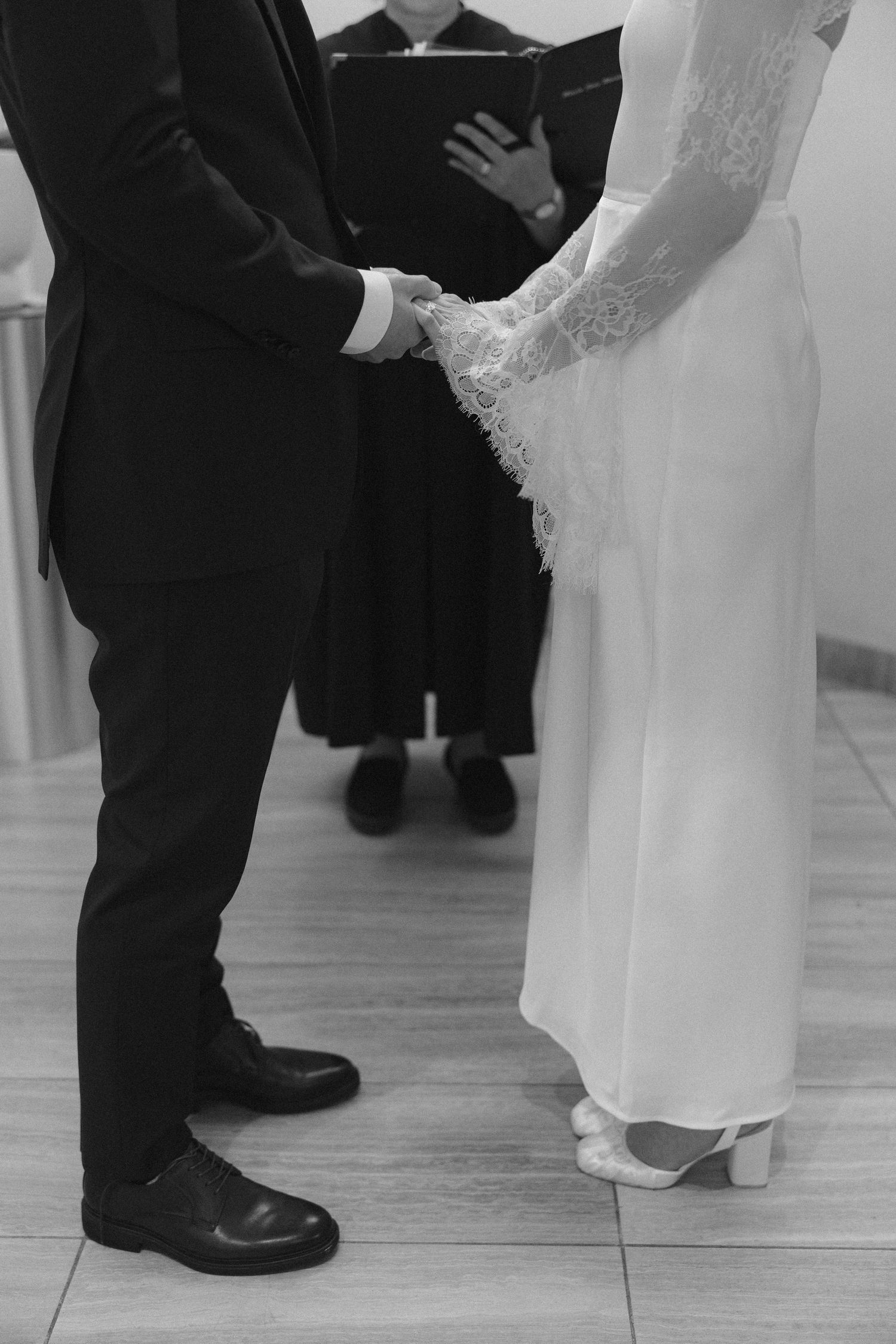 Black and white photo of a bride and groom holding hands during their wedding ceremony as an officiant reads behind them.