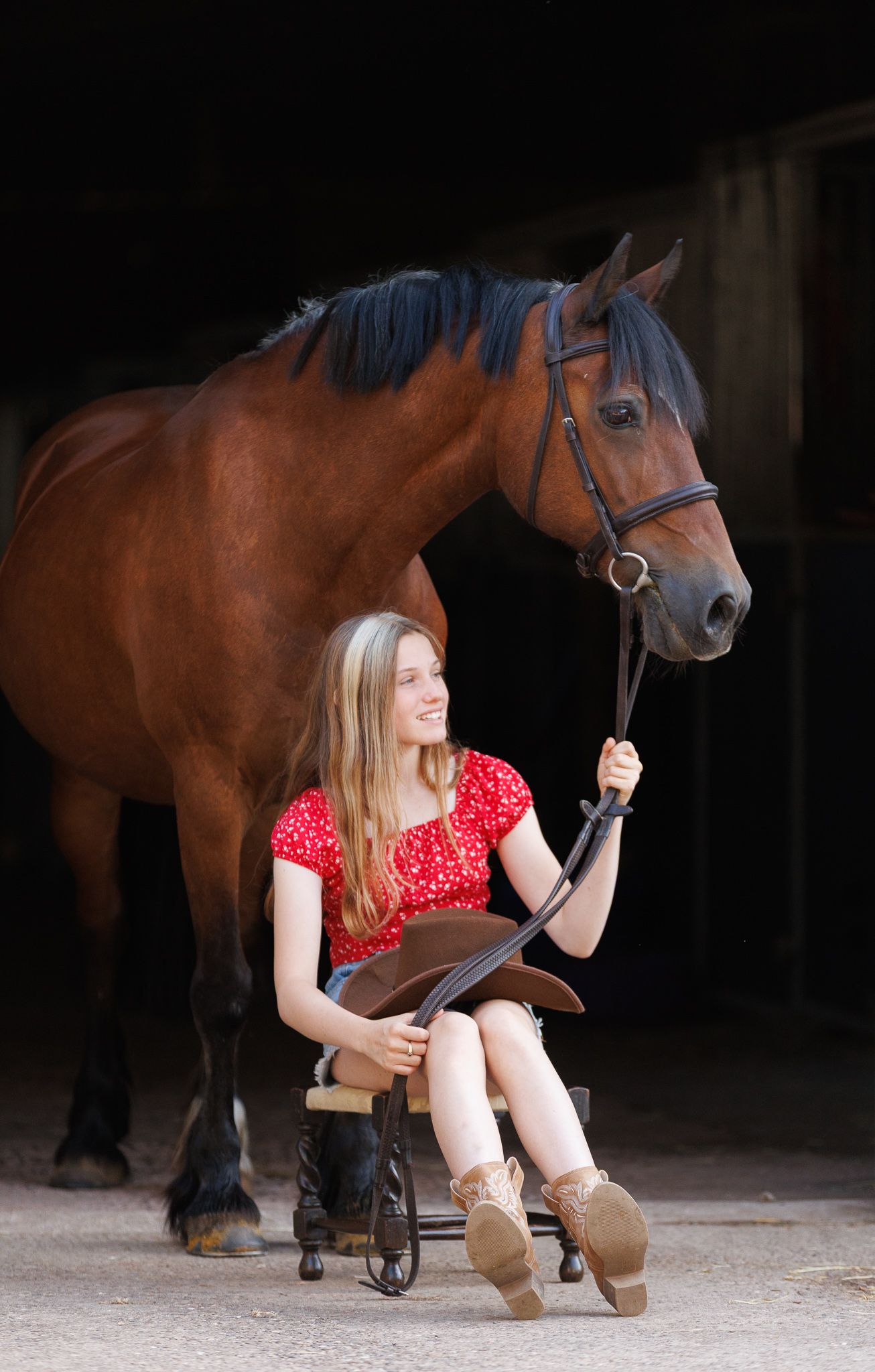 Smiling young woman in a red top sits on a small stool holding the reins of a bay horse in a dark stable, with a brown hat in her lap.