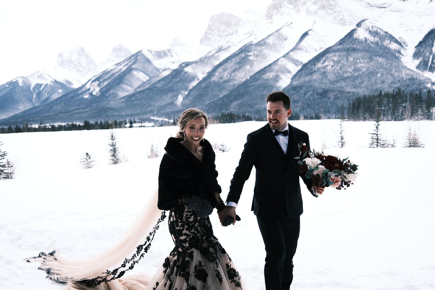 Bride in a fur stole and black lace gown walking hand in hand with the groom through deep snow, veil trailing