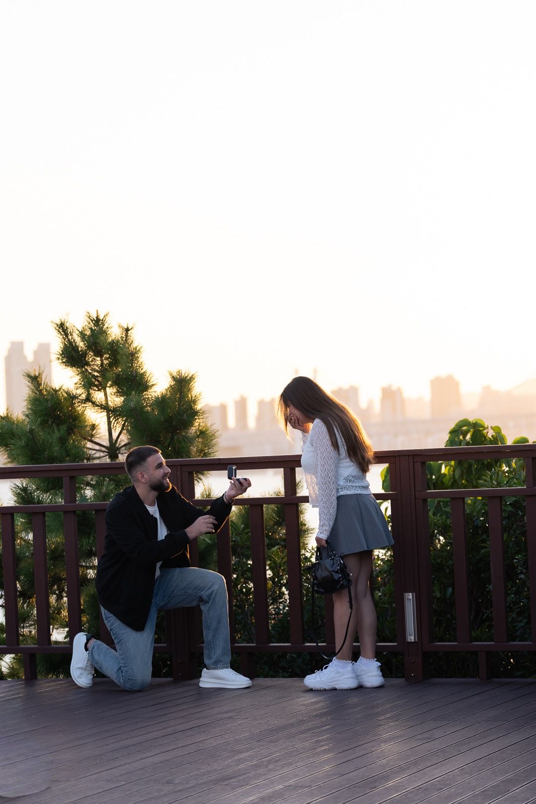 Man on one knee proposing to woman on wooden deck at sunset with city skyline in background.