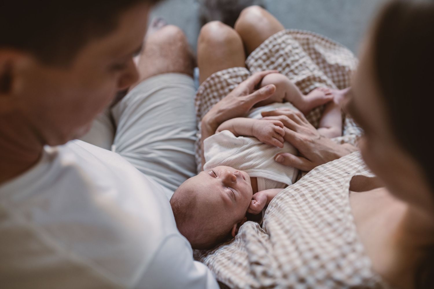 Young couple sitting on bed with newborn baby in modern bedroom with sage green accent wall.