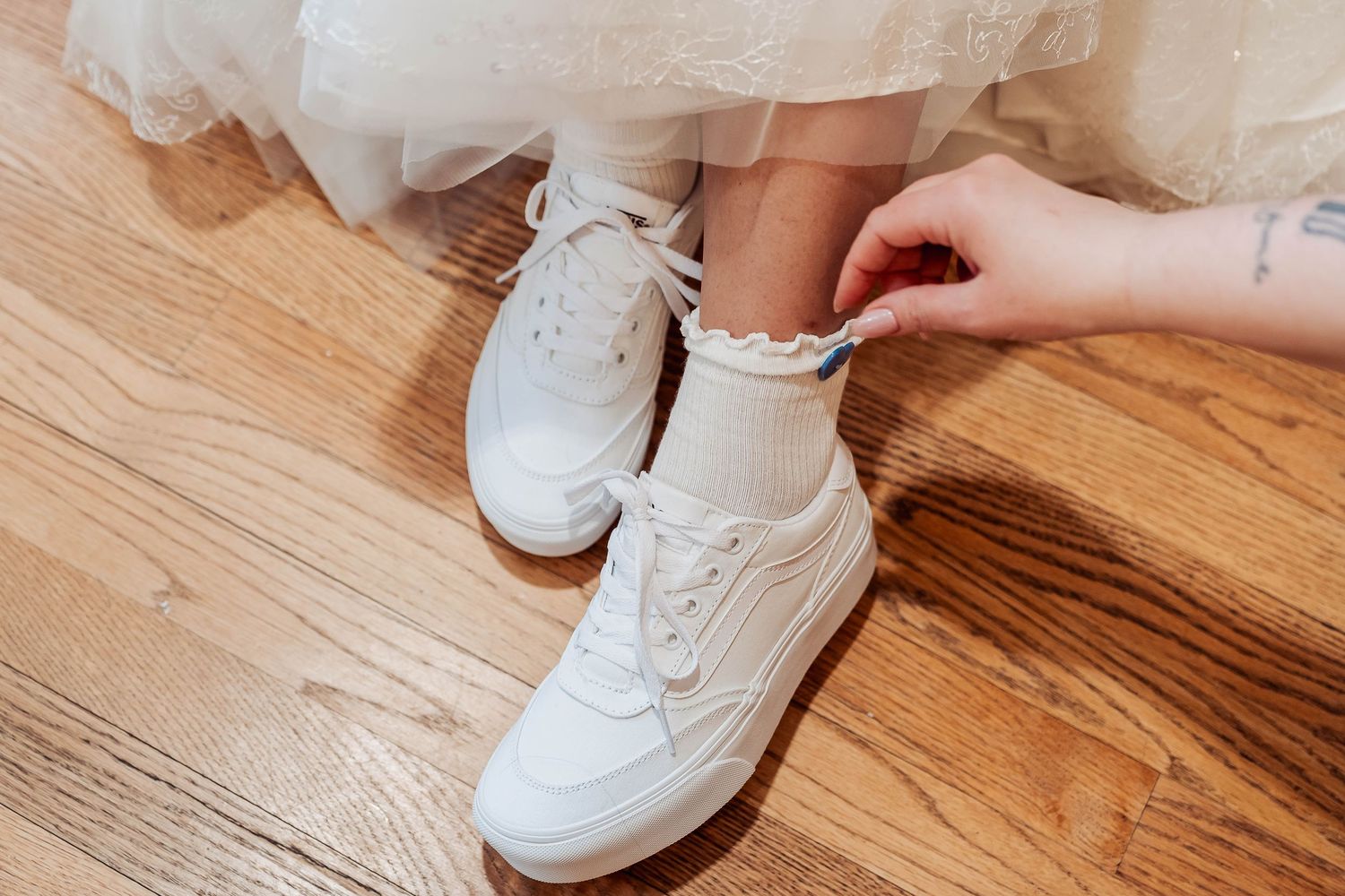 Close-up of white sneakers with ruffled socks being tied, worn under a white tulle dress on hardwood floor.