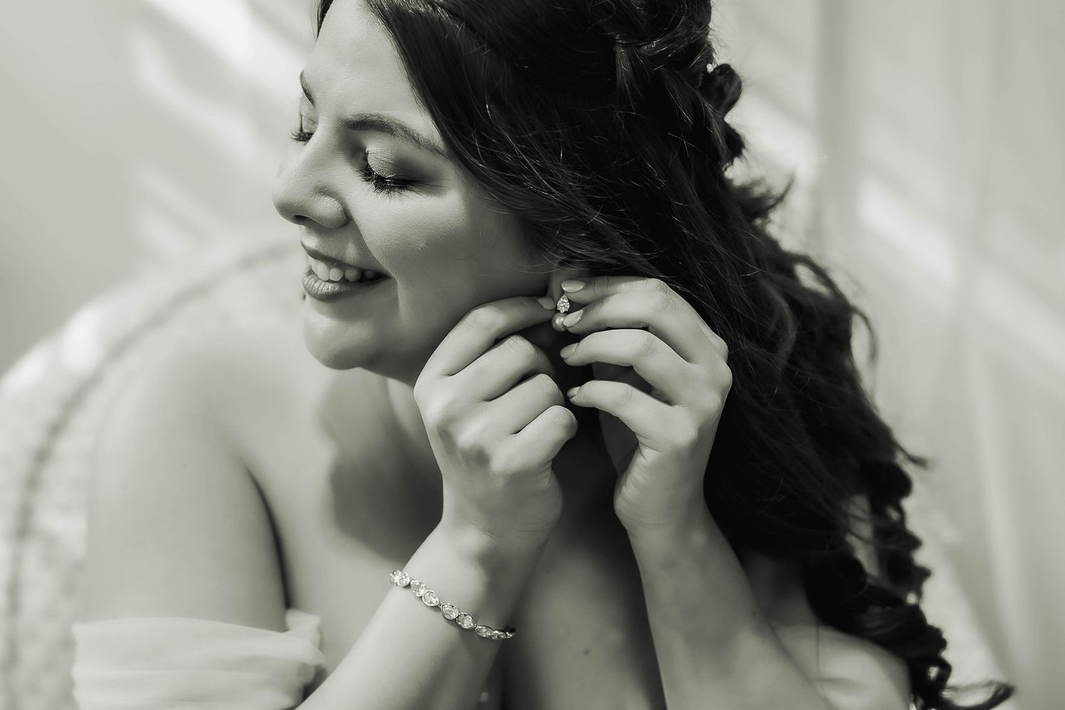 Black and white photo of a smiling bride putting on earrings on her wedding day.