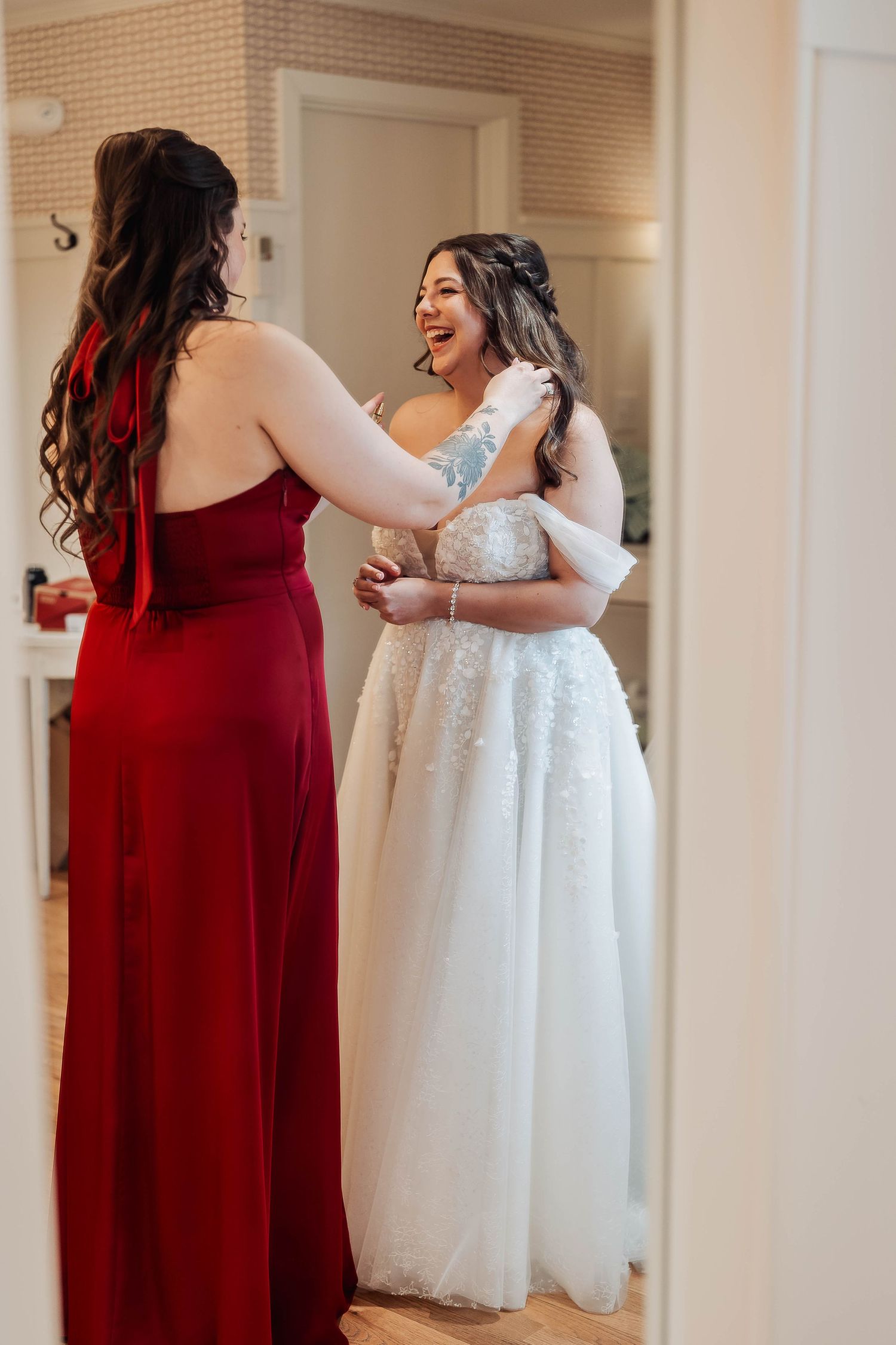 Bride in white gown laughing with bridesmaid in red dress while getting ready for wedding ceremony.