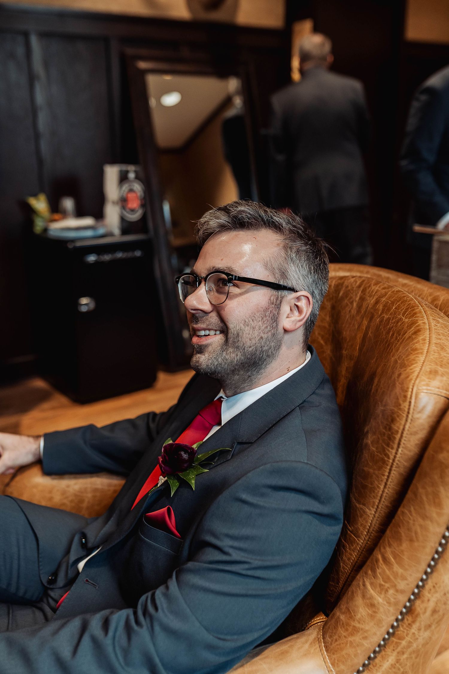 Smiling groom in dark suit with red boutonniere and glasses relaxing in leather chair before wedding ceremony.
