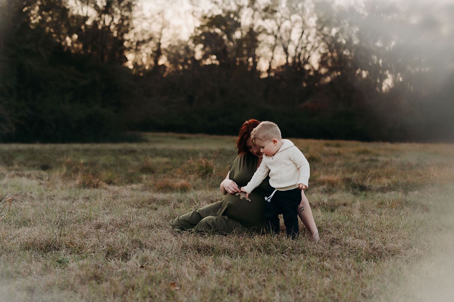 A mother and her young child sit together on a grassy field at golden hour with trees silhouetted in the background.