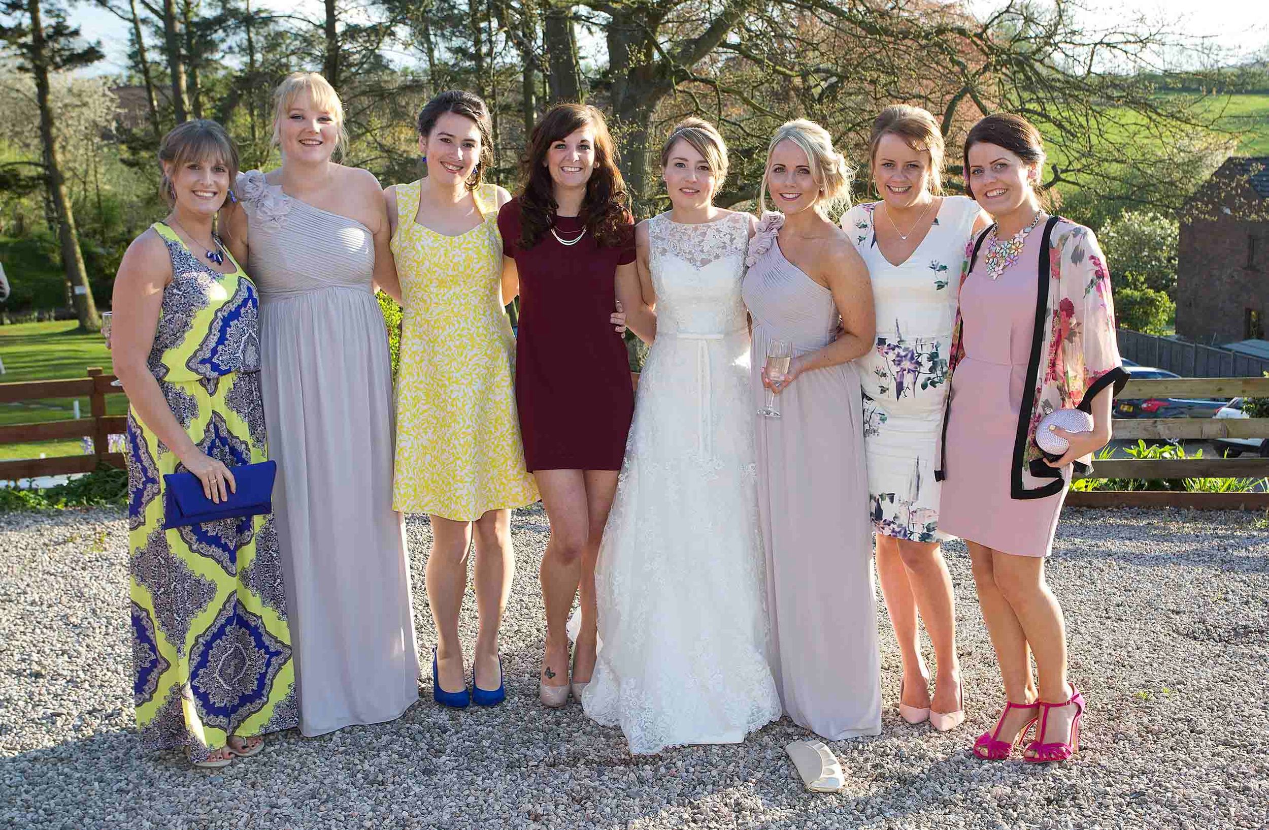 Bride posing outdoors with a group of wedding guests in colourful dresses, with trees and a wooden fence behind them.