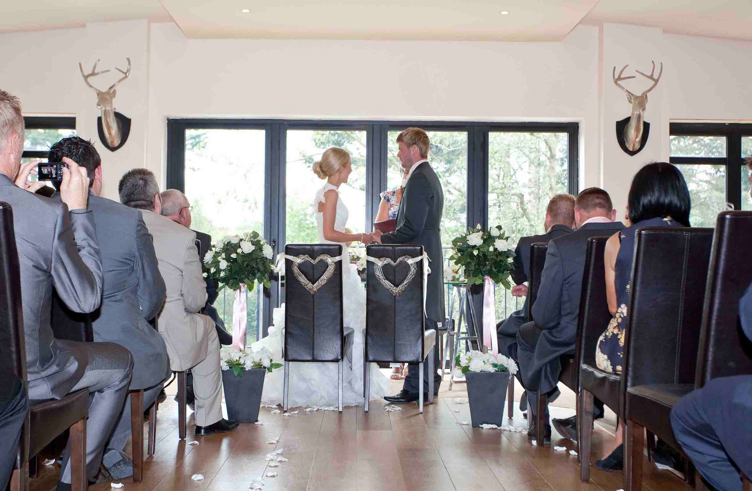 Bride and groom exchanging vows at Heather Glen Country House Hotel, seen down the aisle with seated guests and floral decorations.