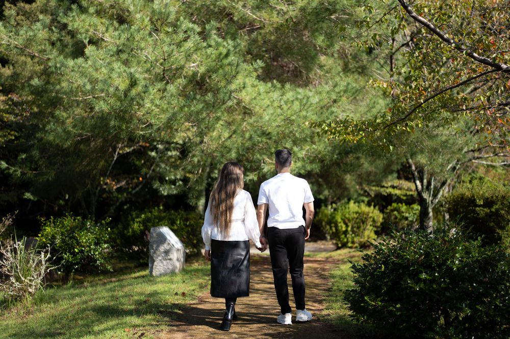Two people in white shirts and dark bottoms walk along a dirt path through a lush green garden with pine trees and shrubs.