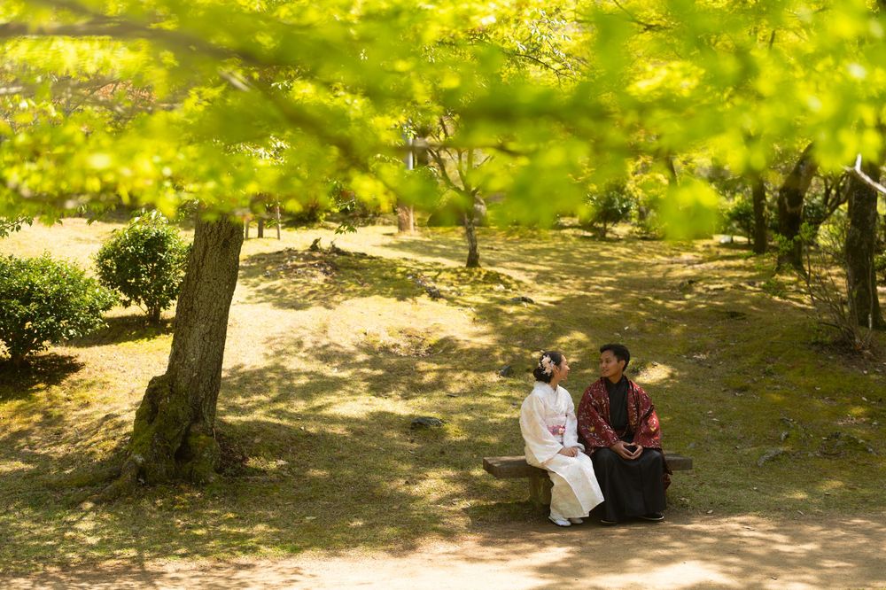 Couple in traditional Japanese attire sit on a wooden bench beneath sunlit green maple leaves in a peaceful garden.