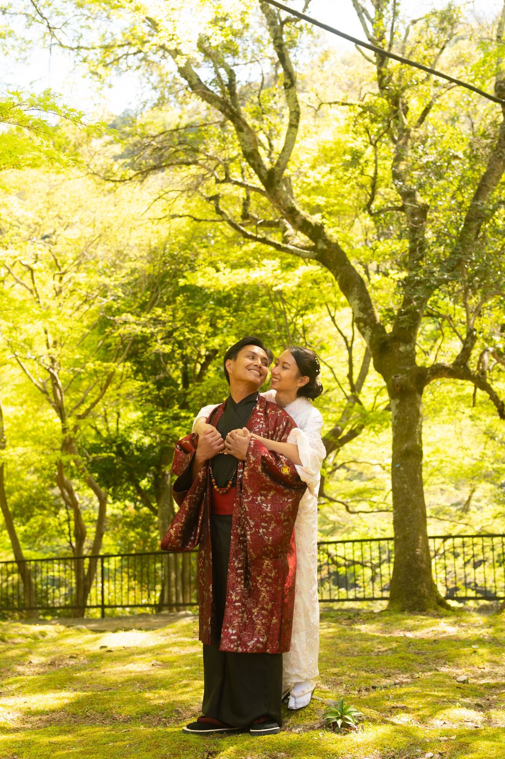 Couple in traditional Japanese attire embracing under green maple trees in a sunlit garden, perfect for a pre-wedding photoshoot.