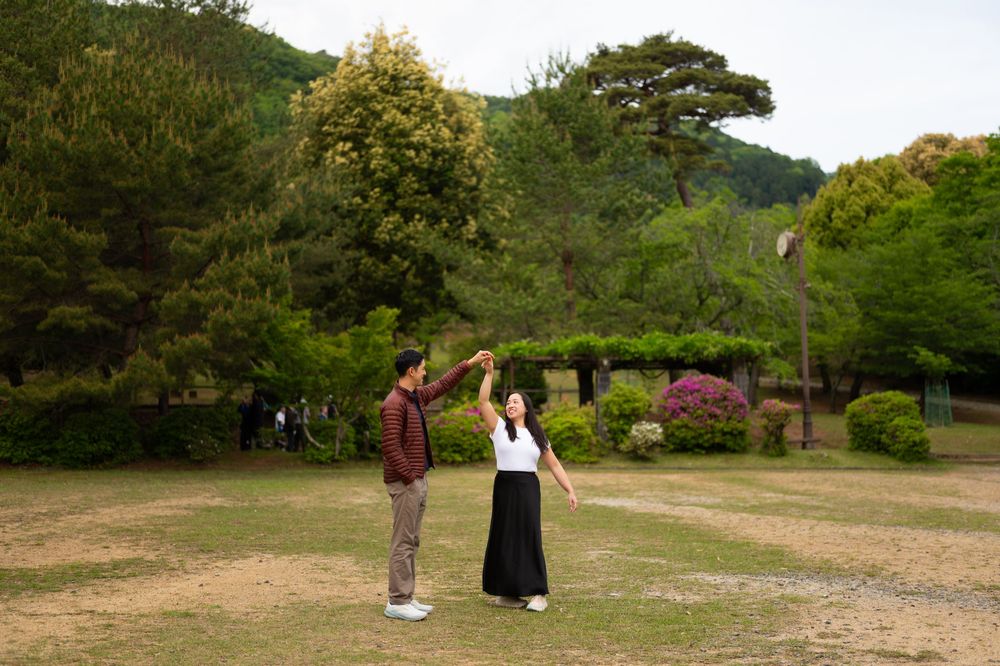 Couple dancing hand in hand on a grassy lawn, the man twirling the woman in a black skirt, with lush green trees and shrubs behind them.