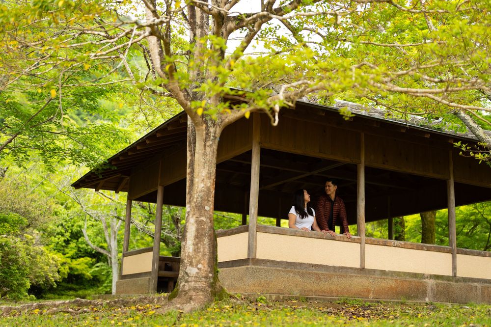 Two people sit together inside an open-air wooden pavilion surrounded by lush green trees in a peaceful Japanese-style garden.