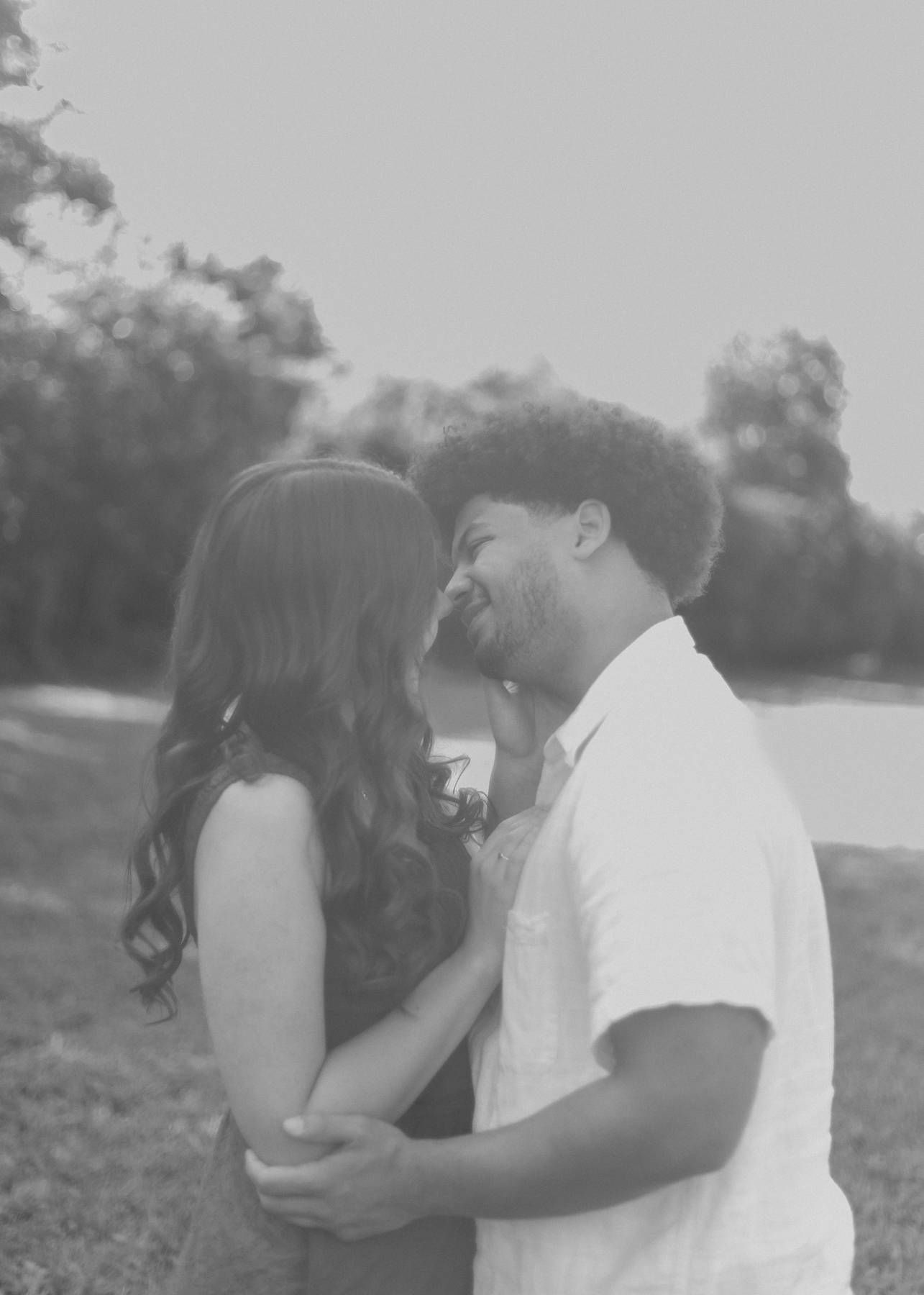 Black and white engagement photo of a couple embracing and smiling near a tree-lined pond.