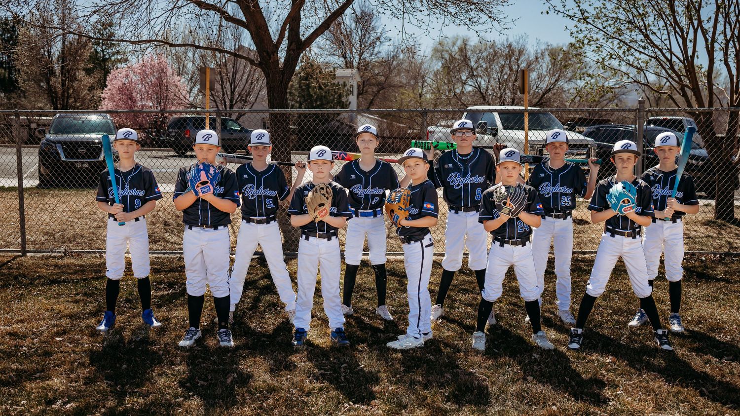 Youth baseball team in matching black jerseys and white pants poses in two rows on a field with trucks and trees behind them.
