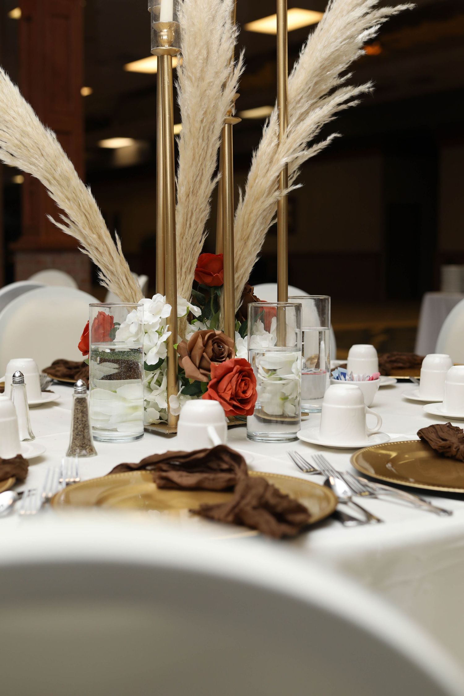 Elegant banquet table centerpiece with pampas grass, rust roses and white flowers beside gold chargers, brown napkins and white teacups.