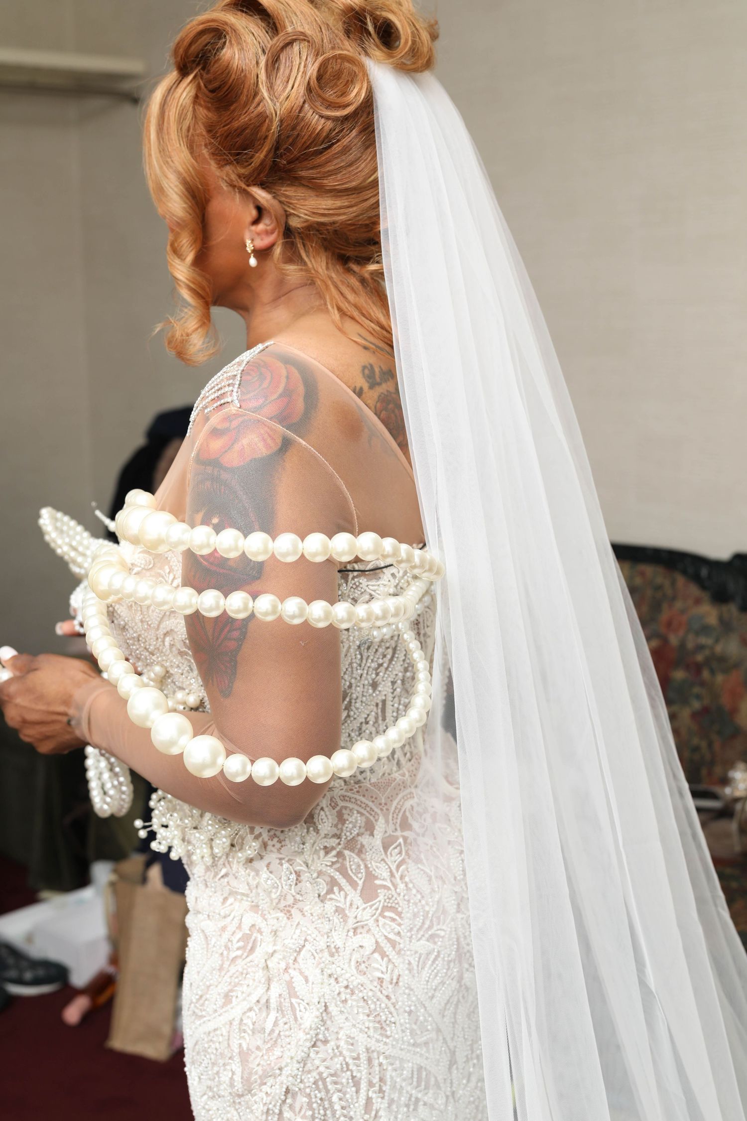 Bride in a lace wedding gown with a sheer back, long veil, floral back tattoo, and layered pearl strands draped over her arms.