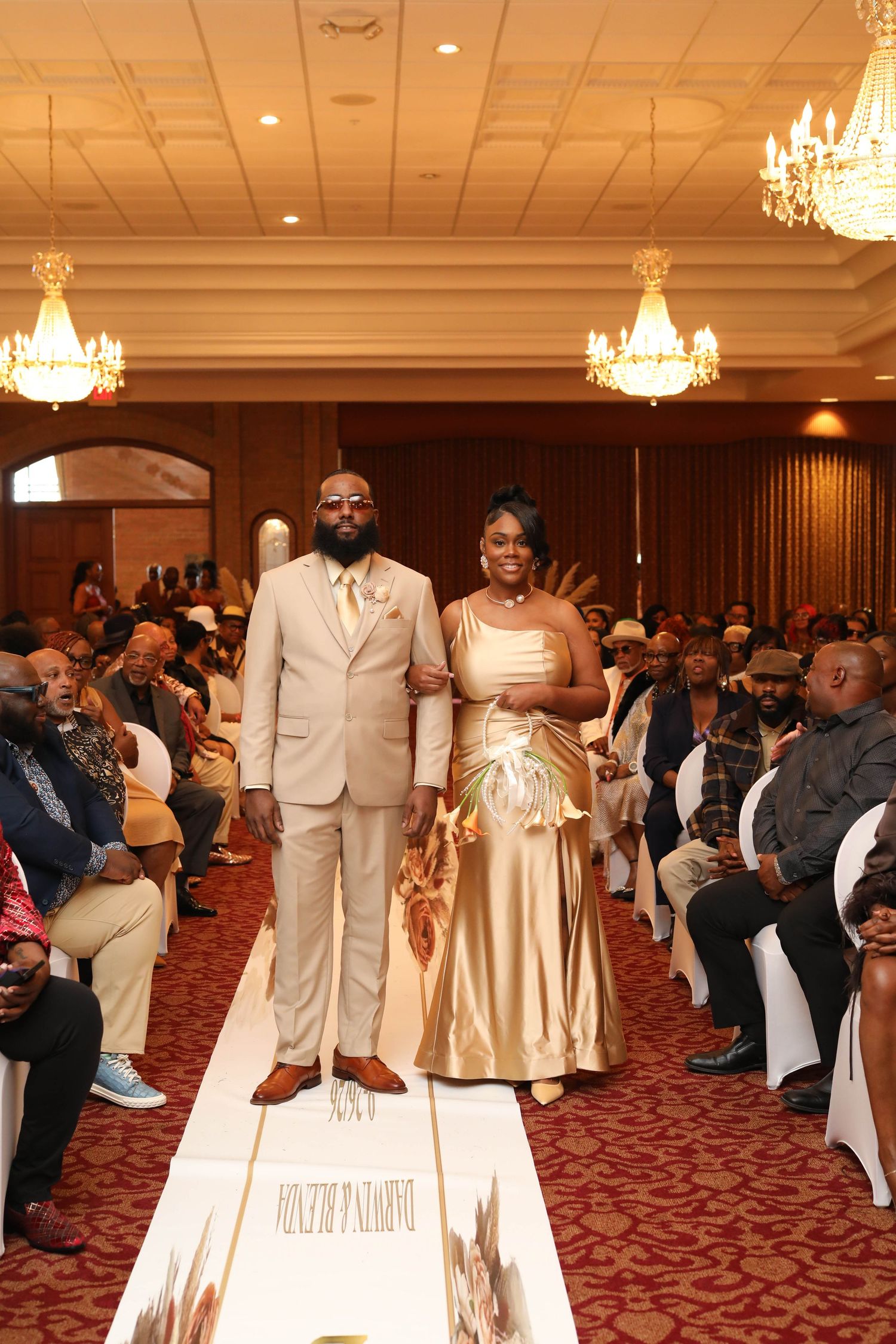 Bride in a gold satin gown and groom in a tan suit walk arm in arm down a white aisle runner past seated guests in an elegant ballroom.