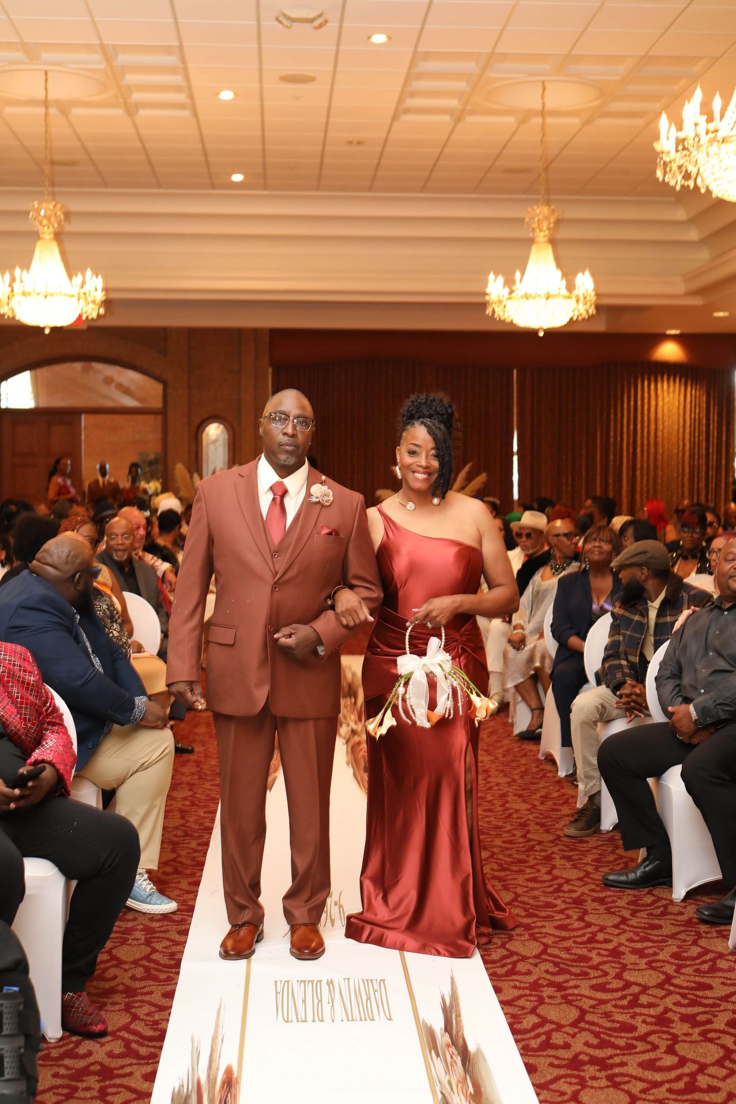 Man in a rust-colored suit escorts a woman in a satin copper gown down the aisle as seated wedding guests look on.
