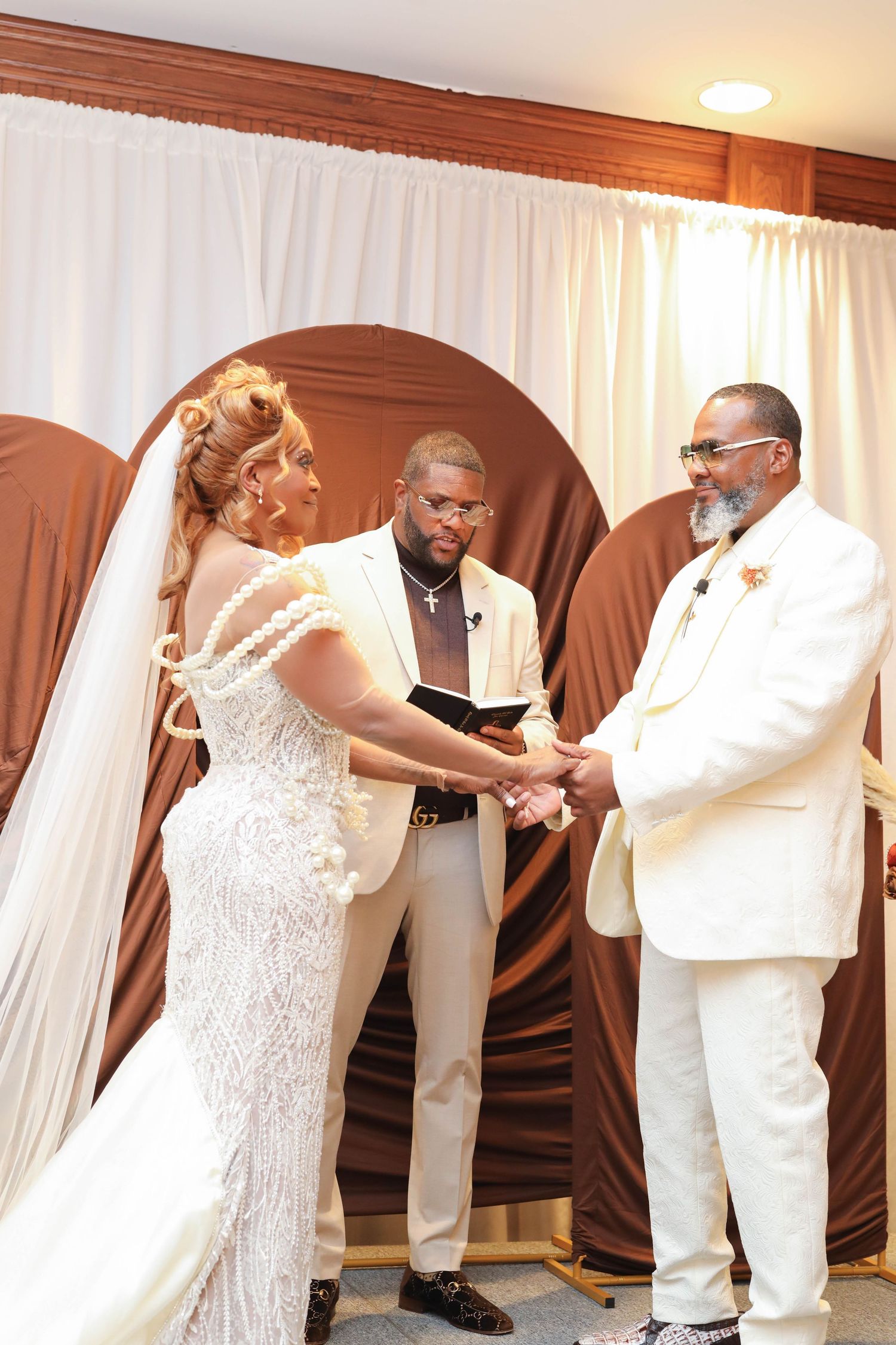 Bride in a beaded lace gown and groom in a white suit hold hands during their wedding ceremony as an officiant reads from a book.