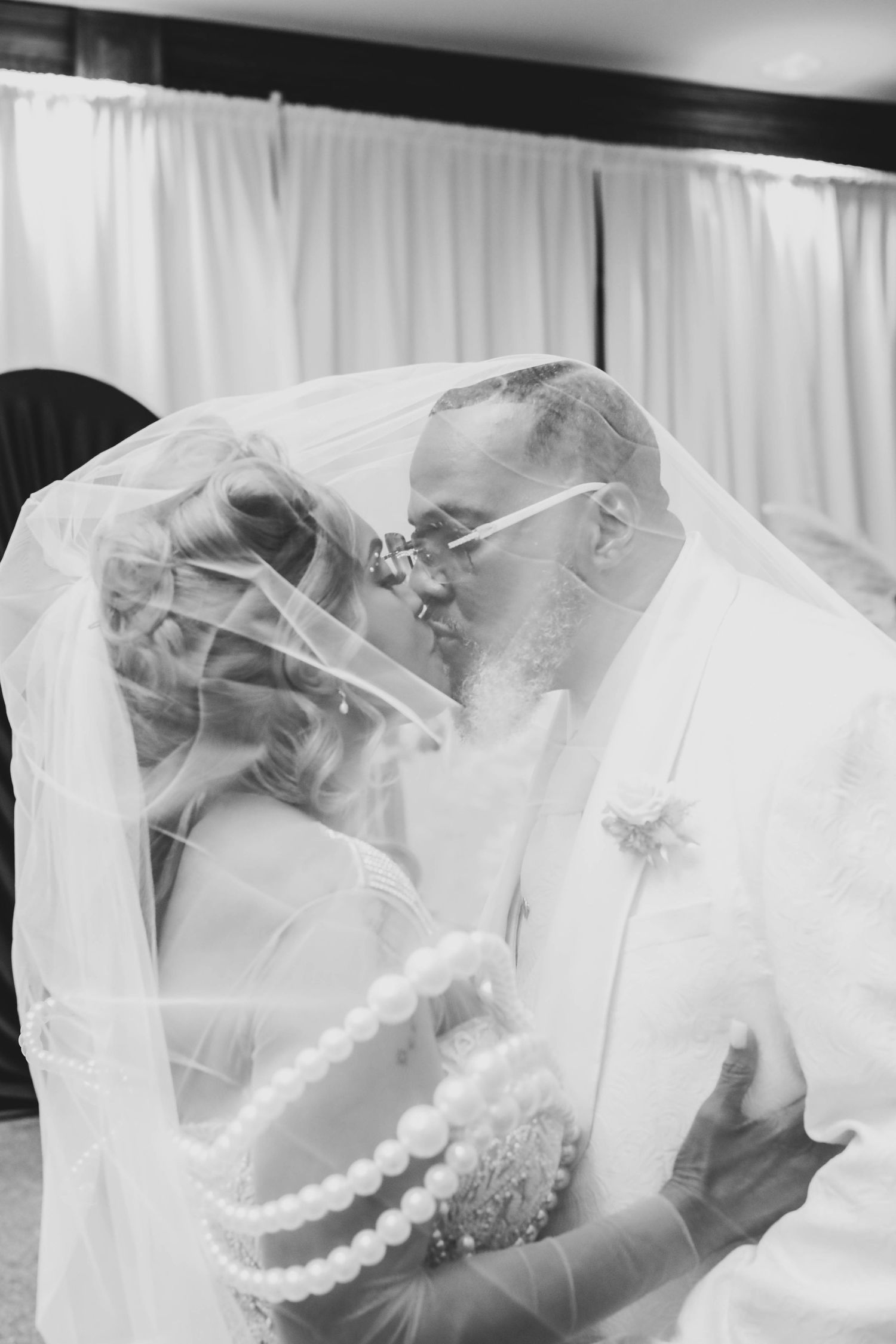 Black and white photo of a bride and groom sharing a kiss under a sheer veil, the bride wearing layered pearl necklaces at their wedding.