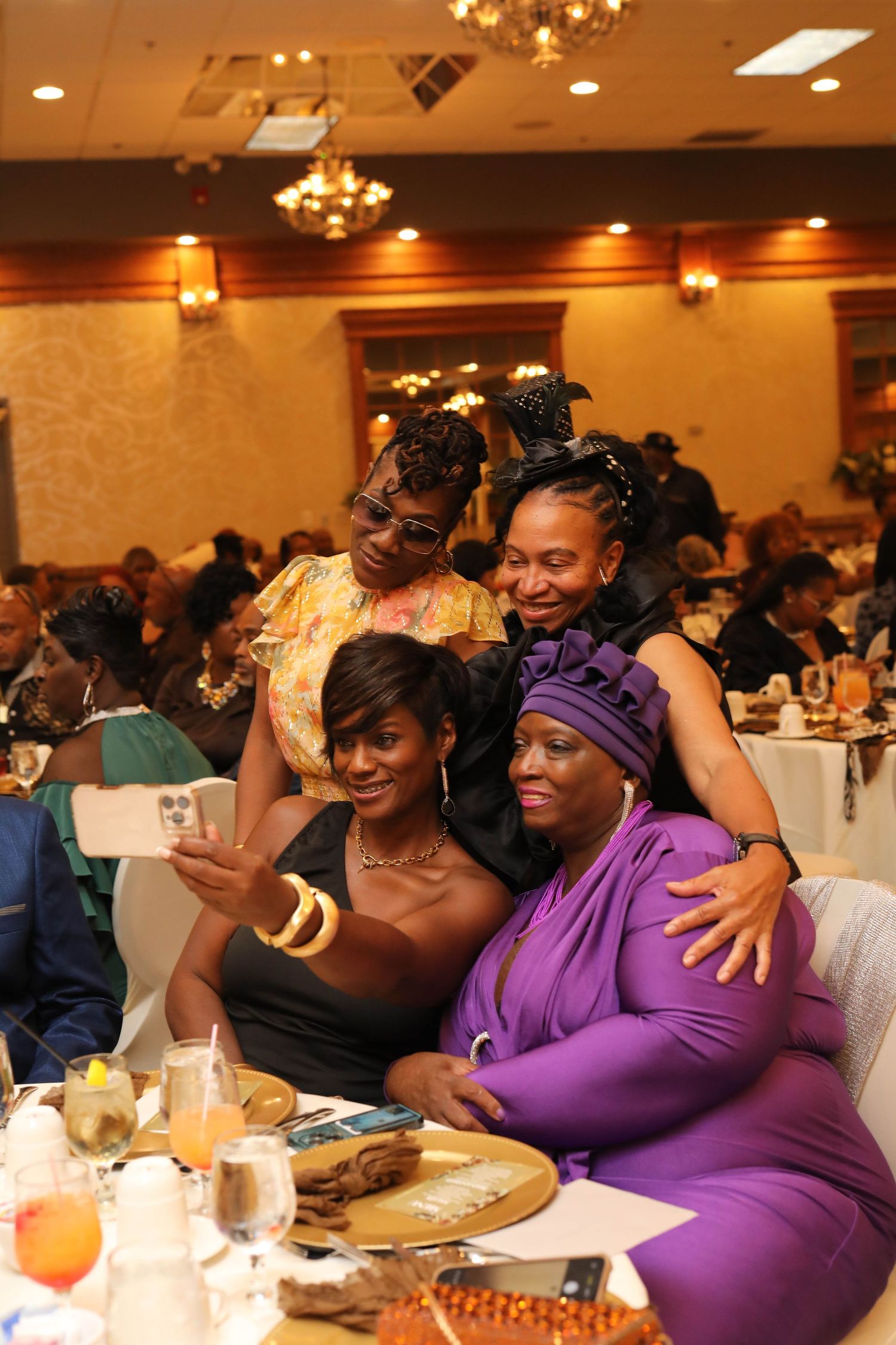 Four smiling women in elegant attire pose for a group selfie at a banquet table set with drinks and plates in a chandelier-lit hall.
